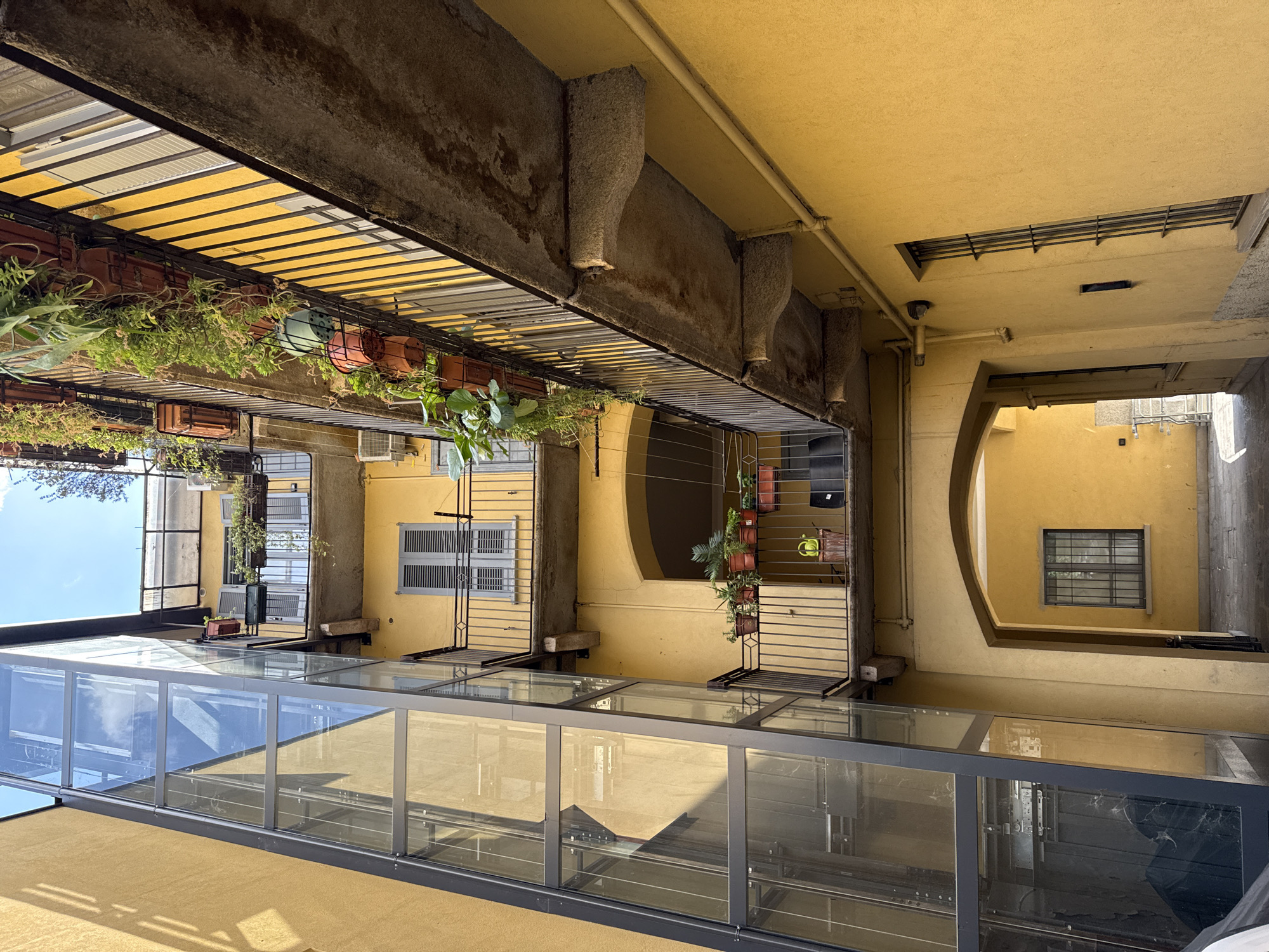The image shows an interior courtyard in Milan, Italy. The perspective is looking upwards, revealing several stories of a yellow building. Balconies are present, some adorned with potted plants. A glass-covered structure is visible at the lower levels of the frame. Windows with gray shutters are spaced intermittently across the walls. The architecture is a blend of modern and older styles. The image appears to be taken during the daytime, with natural light illuminating the scene.