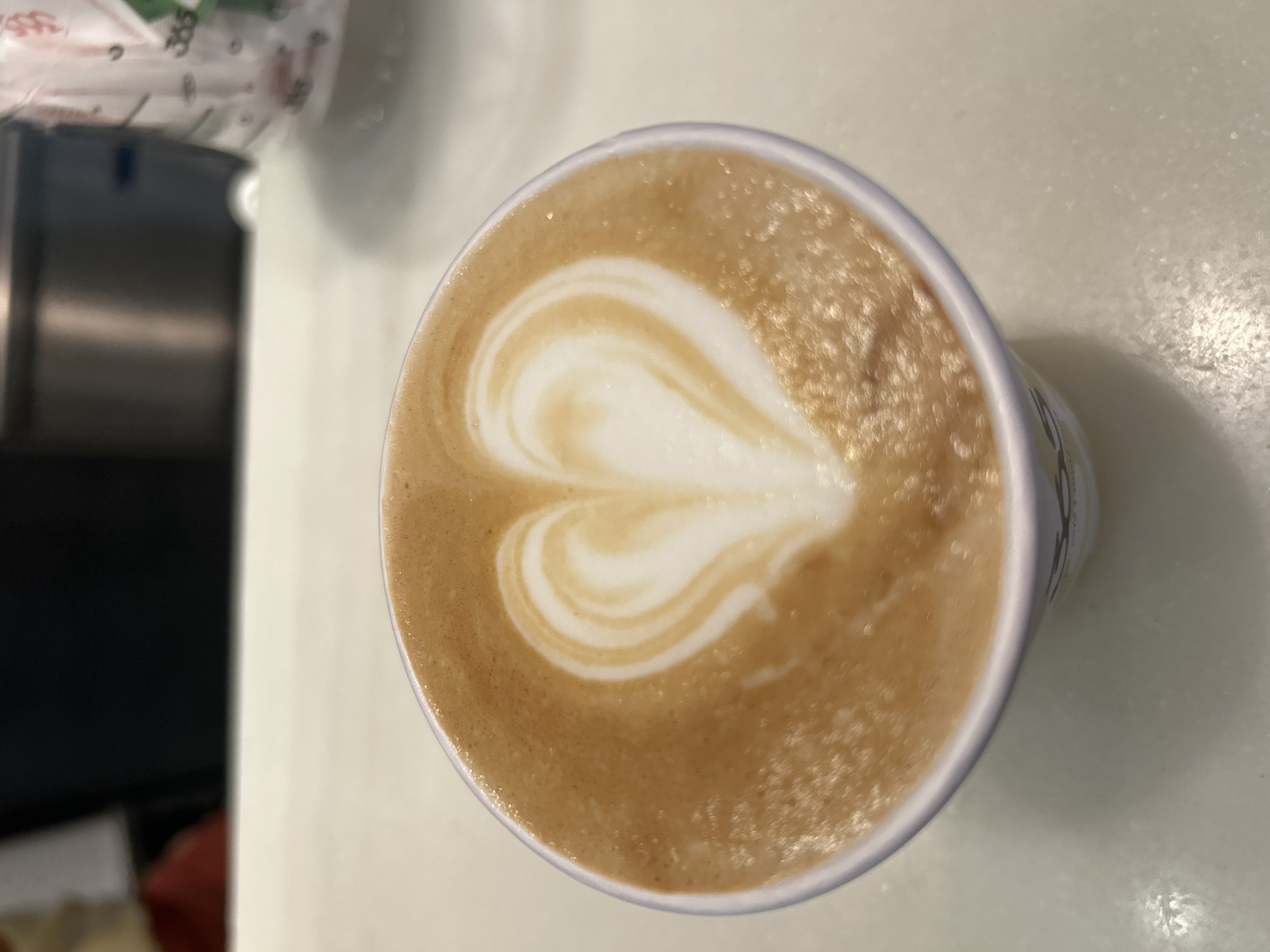 A close-up, high-angle shot shows a disposable white coffee cup filled with latte art. The latte art is in the shape of a heart, with white foam on top of the brown coffee. The cup is on a white, speckled surface, likely a countertop. In the blurred background, there is a dark surface and a partially visible white paper bag with green markings and text. The scene appears to be indoors, possibly in a cafe or a coffee shop. There are no people visible in the image. The lighting is bright, creating some glare on the coffee's surface. The image does not contain any information that would suggest it is located in Barcelona, Spain.
