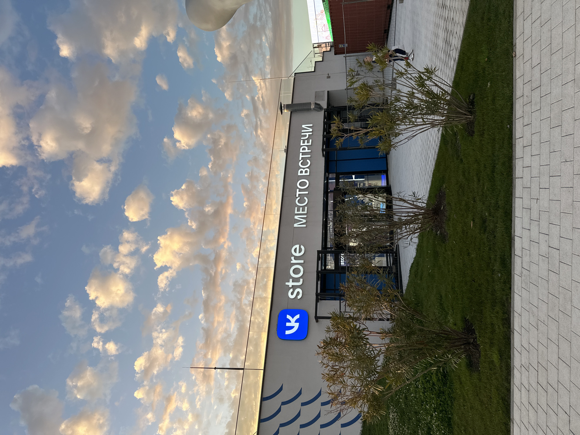 The image depicts a building in Sirius, Russia, identified by a sign that reads "store МЕСТО ВСТРЕЧИ" (Meeting Place). The building's facade is grey with a blue logo of VK (VKontakte, a Russian social media and online social networking service) and blue wave-like design at the base. Tall narrow windows give a view into the shop. In front of the building are small trees planted in a grassy area, bordering a grey paved walkway. The sky in the background is blue with scattered clouds, suggesting either early morning or late afternoon light. No individuals are clearly visible interacting with the building or its surroundings.