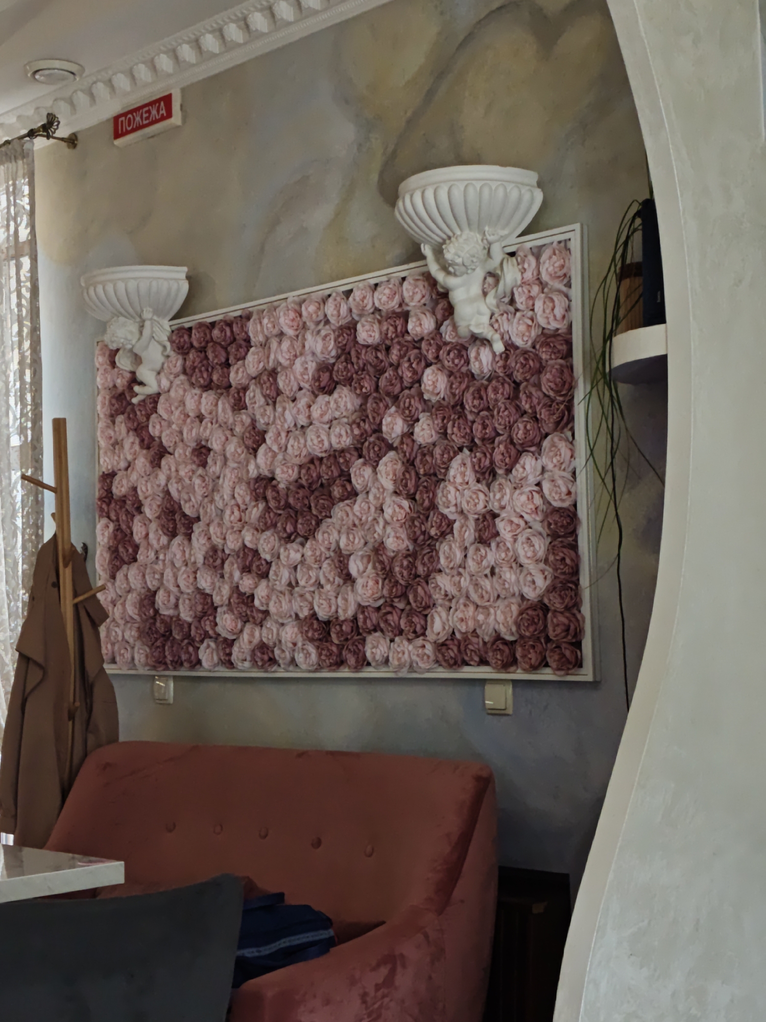 The interior of a room in Vinnytsia, Ukraine, is seen with a pink-reddish couch with buttons along the backrest, positioned in front of a wall adorned with a large, rectangular floral arrangement of pink and burgundy roses, framed in white. The floral arrangement has two small white cherub statues holding white cornucopia-like structures above the corners. A coat rack with a brown jacket stands to the left of the couch. A portion of a white table and a black chair is visible in the foreground. The wall is textured and painted in shades of beige and grey, and there's a small red sign with the Cyrillic word "ПОЖЕЖА" (fire hazard) near the ceiling on the left side. A decorative white architectural element or arch is present on the right side of the frame.