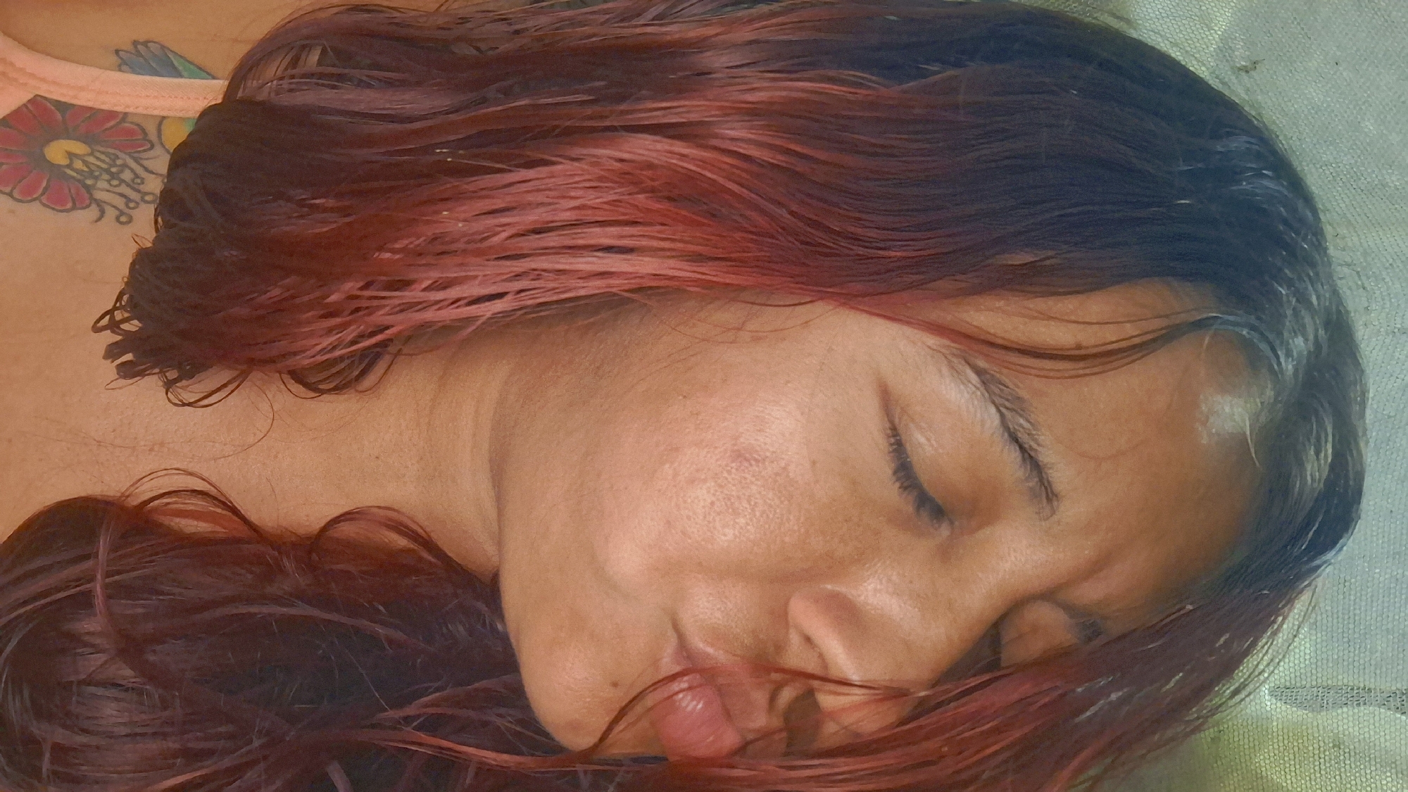 The image is a close-up, high-angle shot of a woman with her eyes closed and lips pursed as if blowing a kiss. Her hair is long, dark, and appears wet, with reddish highlights. She is wearing a light-colored garment, and a colorful tattoo is visible on her upper chest. The background is a neutral, textured surface, possibly a wall or fabric. The scene is indoors, with no indication of an exterior environment or specific location within Vallecito, Venezuela.