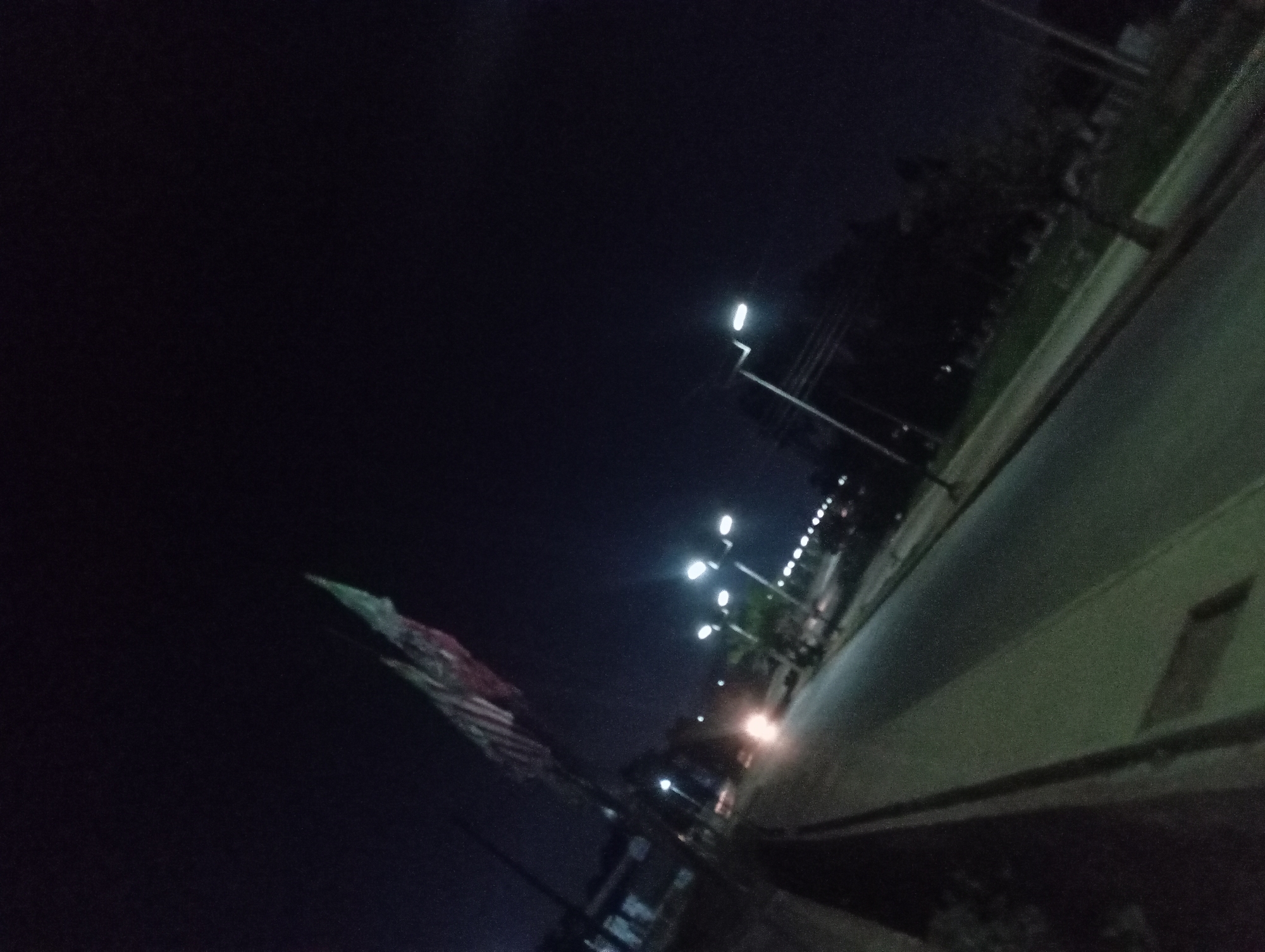 An outdoor, low-light, angled shot captures a street scene at night. A flag, partially illuminated and appearing to be red, white, and green, flutters on a pole on the left side of the frame. The flag is positioned against a dark sky. In the foreground, a paved road runs diagonally across the image, with a raised concrete curb along the left edge. Streetlights with white lights are visible, illuminating the road and surrounding areas. A vehicle's headlights are visible in the distance, creating a bright glow on the road. Trees and buildings are faintly visible in the background, obscured by darkness. The overall impression is of a quiet, urban street at night, possibly in an area with greenery.