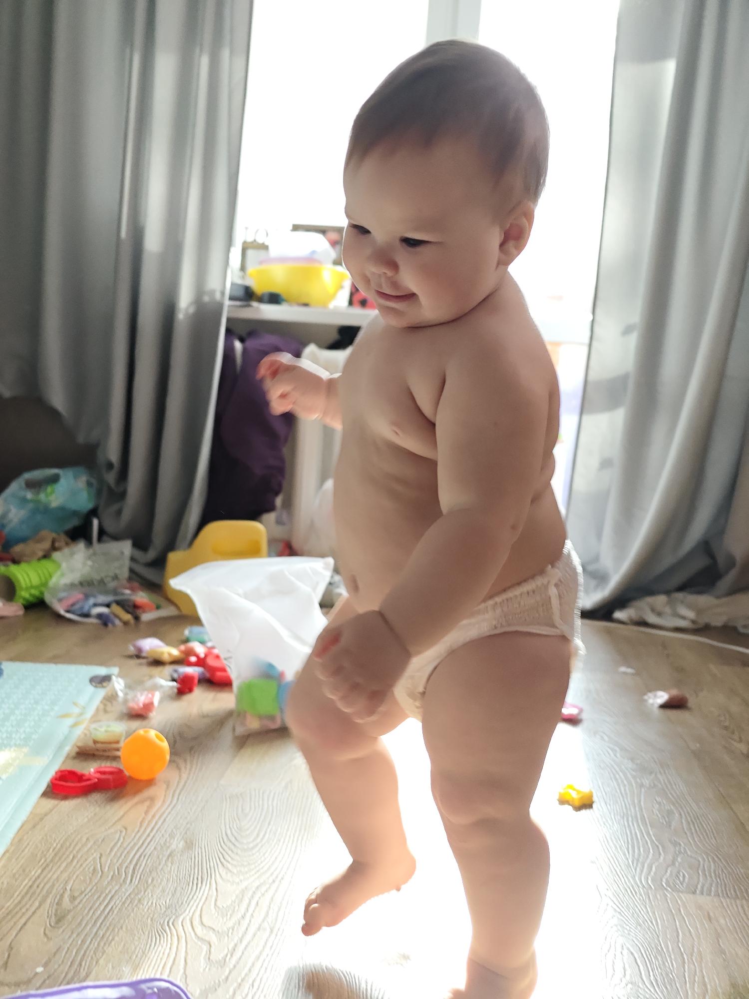 A digital photograph depicts a bare-chested infant, identified as a toddler, standing upright on a light wood laminate floor. The toddler wears a white disposable pull-up diaper and has short, light brown hair. The subject's head is turned slightly to the left, with a gentle smile visible as they look downwards. The body posture suggests motion, with the left leg slightly forward as if taking a step. The environment is an indoor residential setting in Chaikovsky, Russia. The floor is cluttered with various children's toys, including an orange plastic ball, red plastic shapes, multicolored building blocks, and a light blue and green playmat. The background features drawn grey curtains on the left, partially obscuring a window, with a blue plastic bag and a green foam roller visible behind them. To the right, a brightly lit window provides strong backlighting, partially covered by sheer white curtains or blinds. A shelf unit is present against the wall, holding a yellow bowl, purple fabric, and other household items.