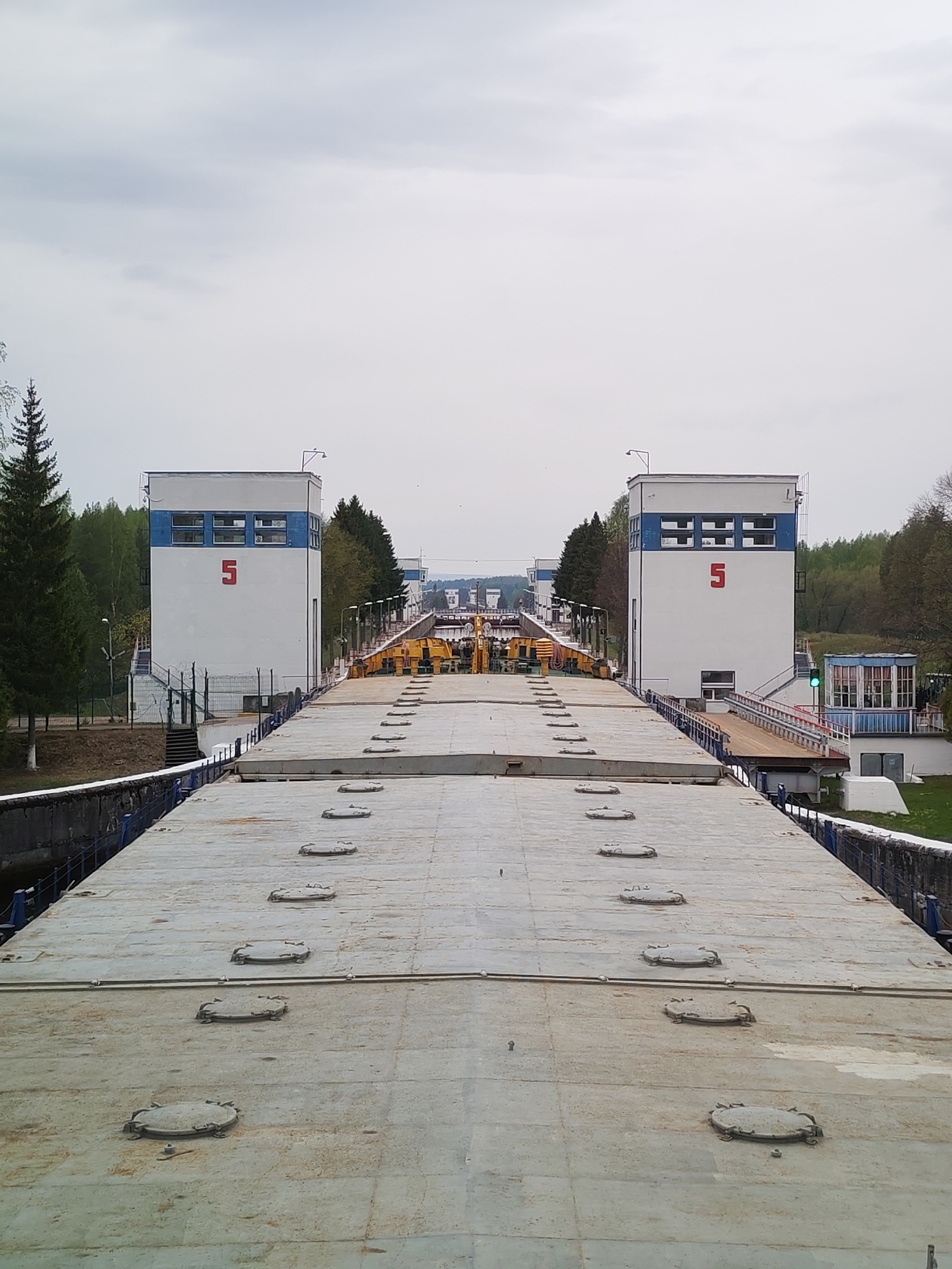The image shows a canal lock, likely the 5th lock in a series. Two symmetrical, white buildings marked with a large red "5" stand on either side of the canal. A concrete bridge or platform with several round hatches stretches across the foreground, providing a high-angle view into the lock system. Various pieces of machinery and equipment are visible between the buildings. Trees and greenery surround the area, suggesting a location in a natural or park-like setting. The sky is overcast. The city of Alekseevskoe is not discernible from the image alone.