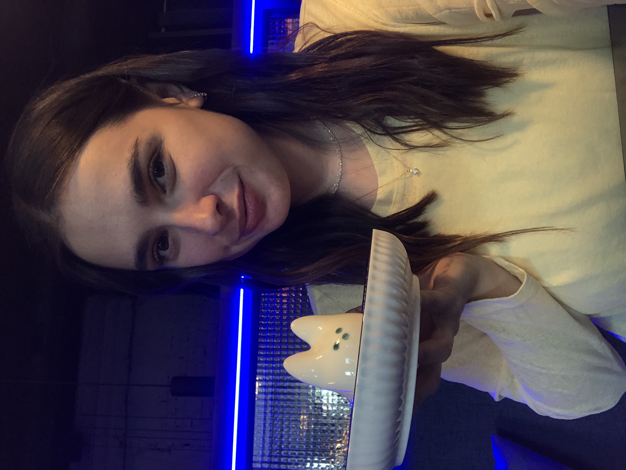 A young woman with long, dark brown hair, wearing a yellow long-sleeved shirt and a delicate necklace, holds a white ceramic dish with a white cat-shaped figurine on it. The figurine has green eyes. The background is dark, with prominent blue neon lighting strips. The overall ambiance suggests an indoor setting, possibly a cafe or restaurant. There is no visible indication of the city of Izhevsk, Russia.