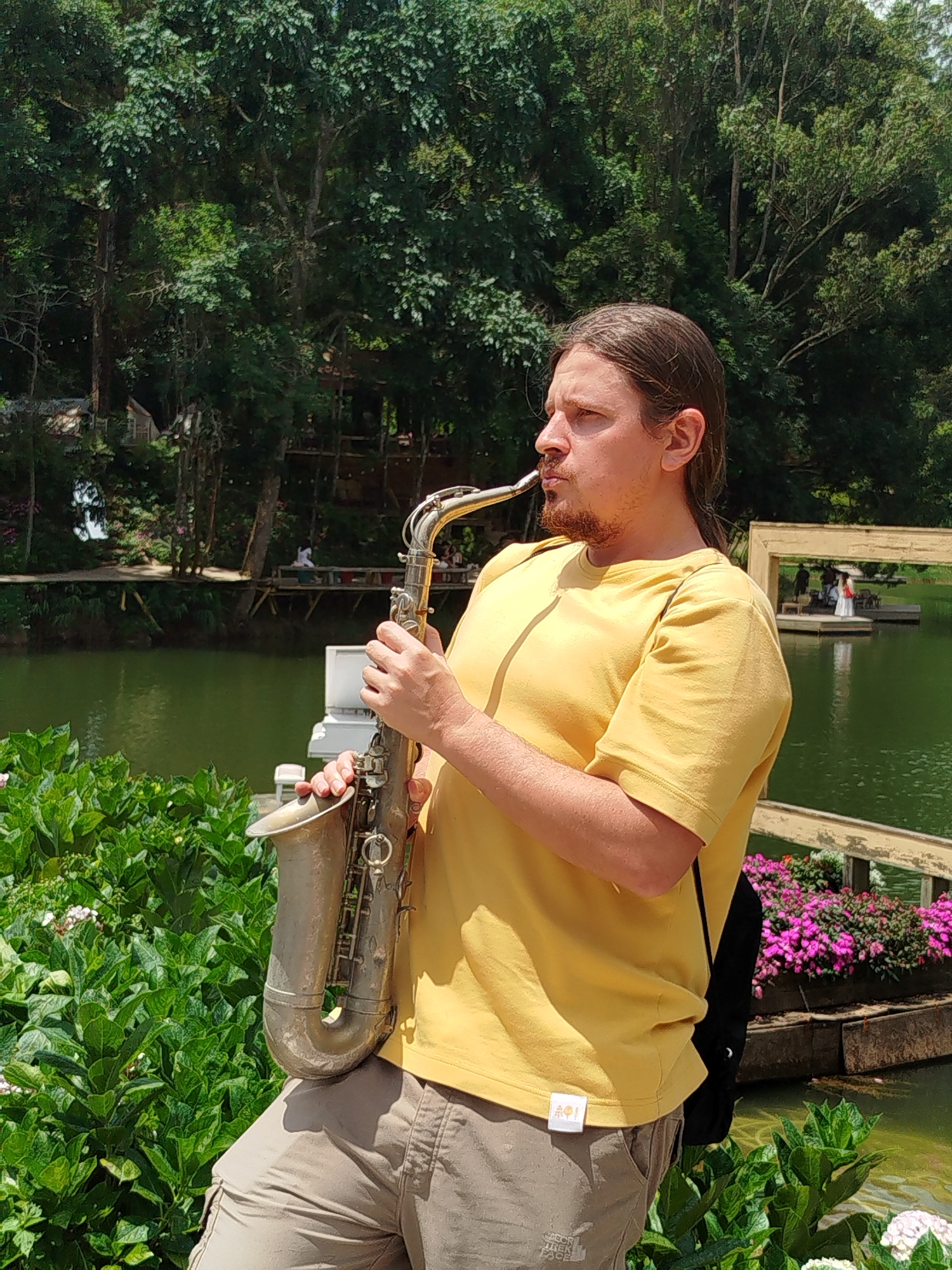 A Caucasian man with long brown hair and a goatee plays a saxophone outdoors. He is wearing a yellow t-shirt and tan cargo shorts. The man is positioned in the foreground, with a body of water and lush greenery behind him. In the background, there are trees, a wooden boardwalk with people, and a small building. The overall environment suggests a park or natural area. The image appears to be taken during the day, with bright lighting.