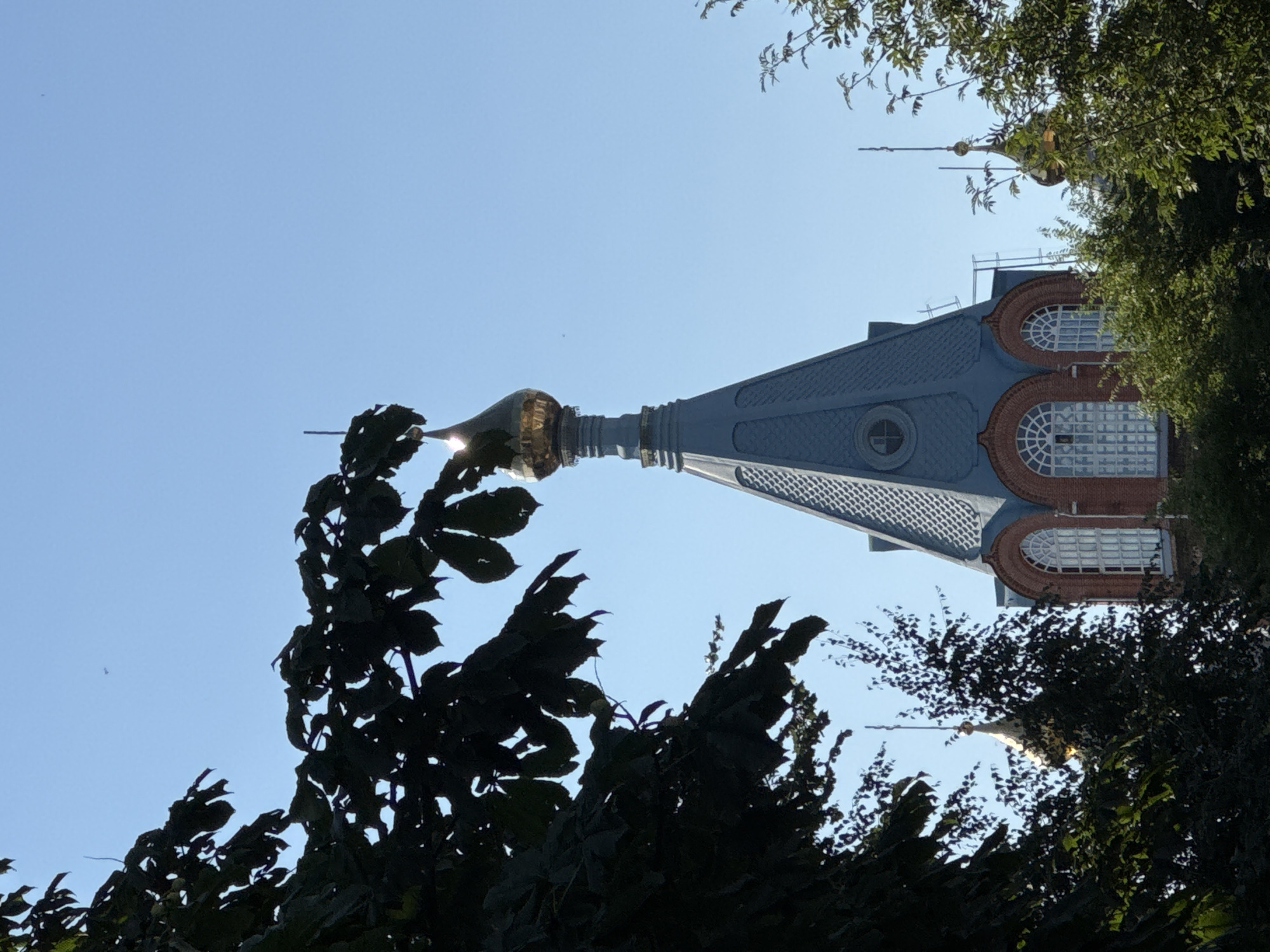 The image shows a clear blue sky above the golden dome of a church tower, which is partially obscured by the dark green leaves of a tree in the foreground. The tower has a grey, conical roof and red brickwork. White arched windows are visible on the tower. In the background, another smaller golden dome is visible, also partially hidden by trees. The scene appears to be a sunny day, with the sunlight glinting off the golden domes. The location is likely a church or monastery in Nizhny Novgorod, Russia, given the architectural style of the domes and towers, which is characteristic of Russian Orthodox churches.