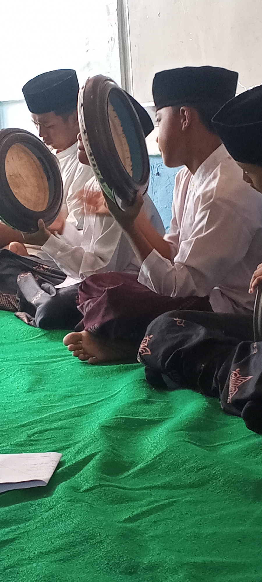The image depicts three young males, likely children or teenagers, engaged in what appears to be a religious or cultural activity. They are seated on a bright green textured surface, possibly a rug or mat. All three are wearing black peci hats, a traditional cap worn by Muslim men and boys. The two individuals in the foreground and mid-ground are holding round, flat percussion instruments, possibly frame drums or tambourines, which are dark in color with lighter rims.
The boy on the left is holding his instrument in front of him, looking down at it. The boy in the middle is holding his instrument to his side, his head turned away from the camera. The third boy, partially visible on the right, is also wearing a peci and a white garment. The clothing worn by the boys is predominantly white, suggesting a formal or religious attire. One boy's lower body is visible, clad in a dark maroon garment. Another boy is wearing socks, one of which has a grey pattern. The environment appears to be indoors, with a light-colored wall and a window visible in the background. The lighting suggests it might be daytime, with natural light entering from the window.
While the image does not explicitly provide location details for Lumajang, Indonesia, the presence of peci hats and the attire strongly suggests a Muslim cultural context common in Indonesia. This scene could be taking place in a mosque, a madrasah (Islamic school), or a private home during a religious gathering or practice session.