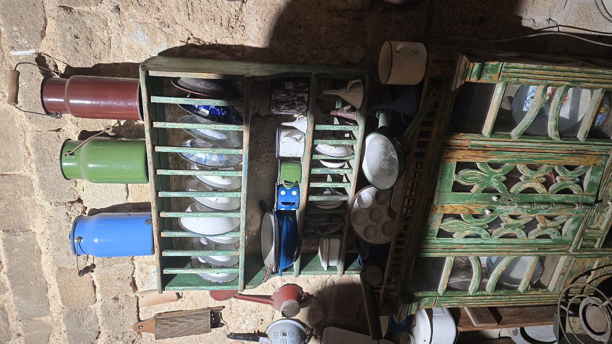 This is a medium shot, eye-level photograph of a rustic kitchen in Acre, Israel. The scene is dominated by an old, green, distressed wooden hutch filled with various kitchenware. The hutch has open shelves above and closed cabinet doors below, with intricate carved detailing. On the top shelf, there are three large, colorful enamel jugs: one blue, one green, and one maroon. Below these, the shelves are filled with plates, bowls, mugs, and kitchen utensils such as strainers and funnels. The kitchen is situated against a rough-hewn stone wall, typical of the historic architecture found in Acre's Old City. The lighting is somewhat dim, with shadows casting across the stone wall and highlighting the textures of the objects. There are no people visible in the image.