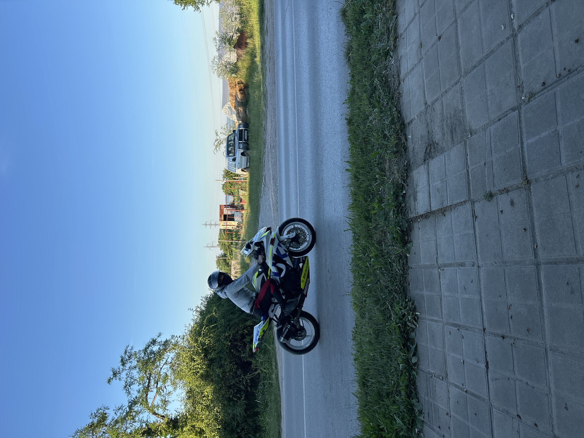 A person wearing a gray hoodie, black helmet, and dark pants rides a white and blue sport motorcycle on a two-lane asphalt road. The motorcycle has yellow accents and the word "DUNLOP" printed on the tire. The rider is positioned on the right side of the road, moving away from the viewer. To the left of the road is a patch of green grass and then dense green trees. In the background, on the right side of the road, there is a white SUV parked on the side, a pile of hay bales, a building under construction with exposed beams, and what appears to be power lines. A brown animal, possibly a cow, is visible grazing in the distance. The sky is clear and blue. The foreground shows a concrete sidewalk made of square pavers.