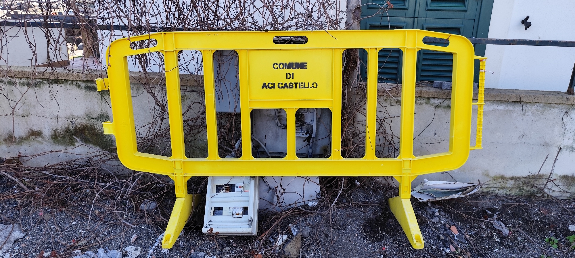 A yellow plastic barrier stands in front of a utility access panel near a building in Aci Castello, Italy. The barrier reads "COMUNE DI ACI CASTELLO" in black lettering. The utility panel is open, exposing its internal components. Debris and leafless branches surround the barrier. The background features a light-colored wall and dark-green shutters.