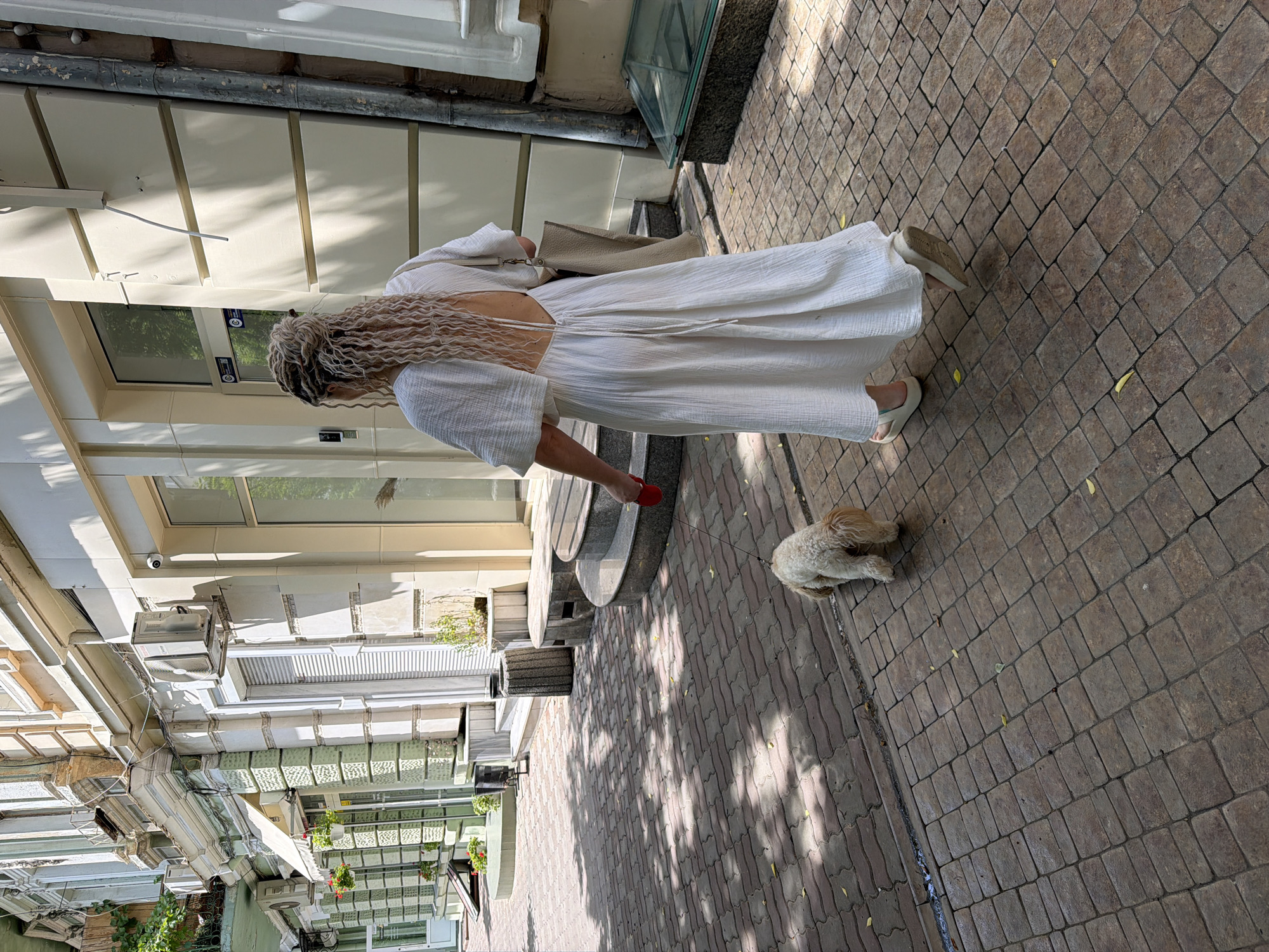 A woman with blonde, curly hair walks away from the camera down a cobblestone sidewalk in Odesa, Ukraine. She is wearing a long, white, open-back dress and light-colored sandals. She carries a light-colored tote bag over her right shoulder and holds a red leash attached to a small, light-colored dog walking ahead of her. The dog appears to be sniffing the ground. Sunlight casts dappled shadows on the sidewalk. The background features the facades of buildings with ornate architectural details, air conditioning units, windows, and doorways. A green planter with red flowers is visible on the left. The scene is a street in a European city, consistent with Odesa's architecture.