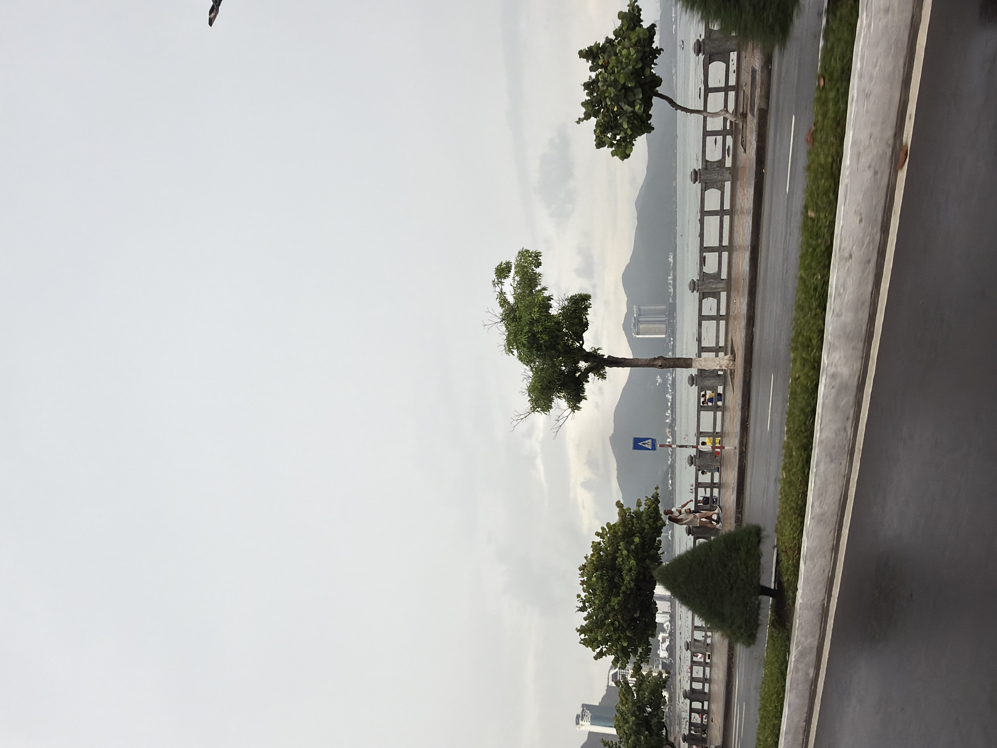 The image captures a street scene in Bac Nha Trang Ward, Vietnam. The foreground shows a road with a concrete barrier on one side, and a pedestrian walkway with trees and a fence on the other. The background reveals a waterfront with mountains in the distance. A few people are visible on the walkway. The sky is overcast.