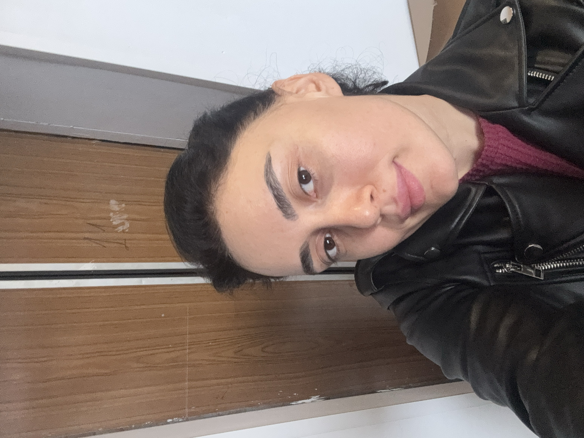 A close-up portrait depicts a female subject from the chest up, centrally positioned and looking directly at the camera with a slight smile. She has dark hair pulled back, dark eyebrows, and brown eyes, with a fair to medium complexion. She is attired in a black leather-style jacket, featuring a visible silver zipper and studs on the collar, layered over a ribbed, dark pink or maroon sweater. The background is an interior setting. To the left of the subject, a brown wooden panel or door with vertical grain is visible, bearing faint white markings, including a possible "11" near the top right. A dark vertical strip separates this wooden surface from a plain white wall on the right. The specific location within Tbilisi, Georgia, cannot be determined from the provided visual information.