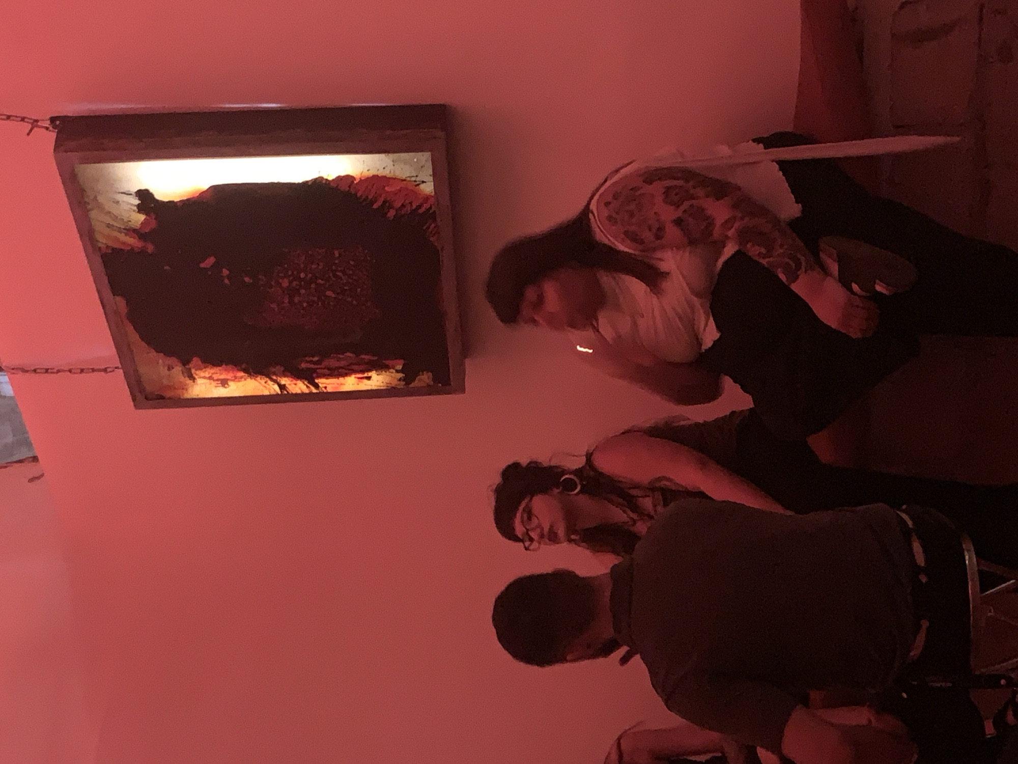 A dimly lit, predominantly red-toned interior space contains a framed, dark-colored artwork hanging on the wall. Beneath the artwork, three individuals are present. One individual, seen from the back, is wearing a dark-colored shirt. Another individual, also facing away from the camera, has dark hair and glasses. The third individual, turned towards the artwork, has tattooed arms and wears a light-colored top with dark pants. No location data is available.