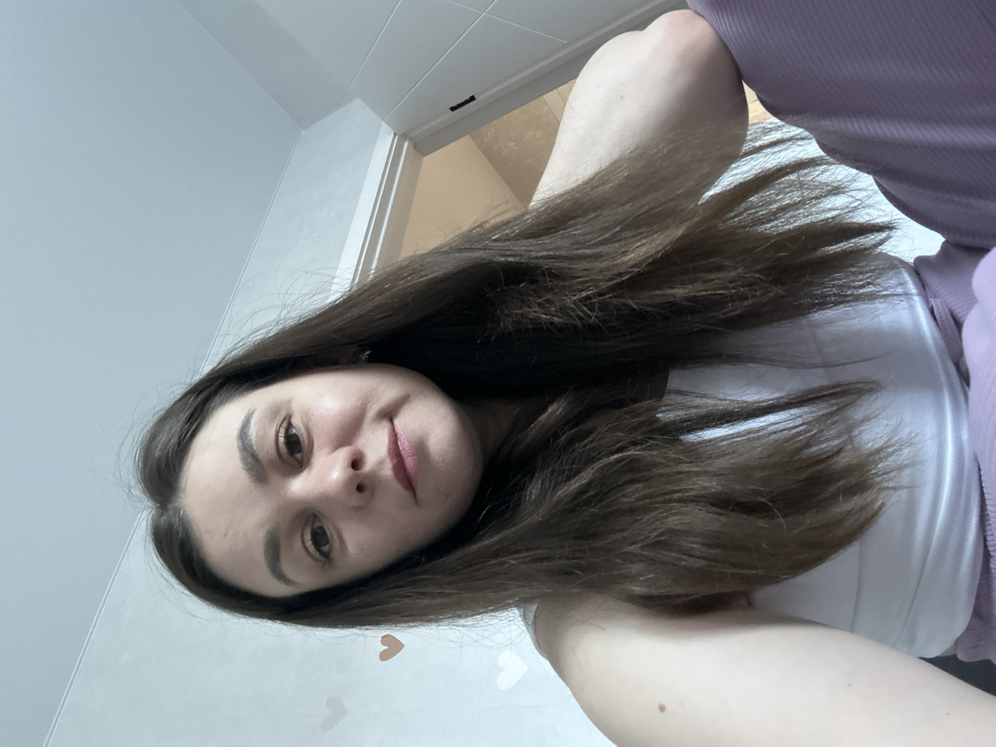 A person with long, dark brown hair is lying down, facing the camera. They are wearing a white top and a light purple garment. The background is a white wall with faint heart-shaped decorations and a glimpse of a tiled room through a partially visible doorway. The location is inferred to be in Chaikovsky, Russia, based on the user's query, but there is no specific evidence in the image to confirm this.