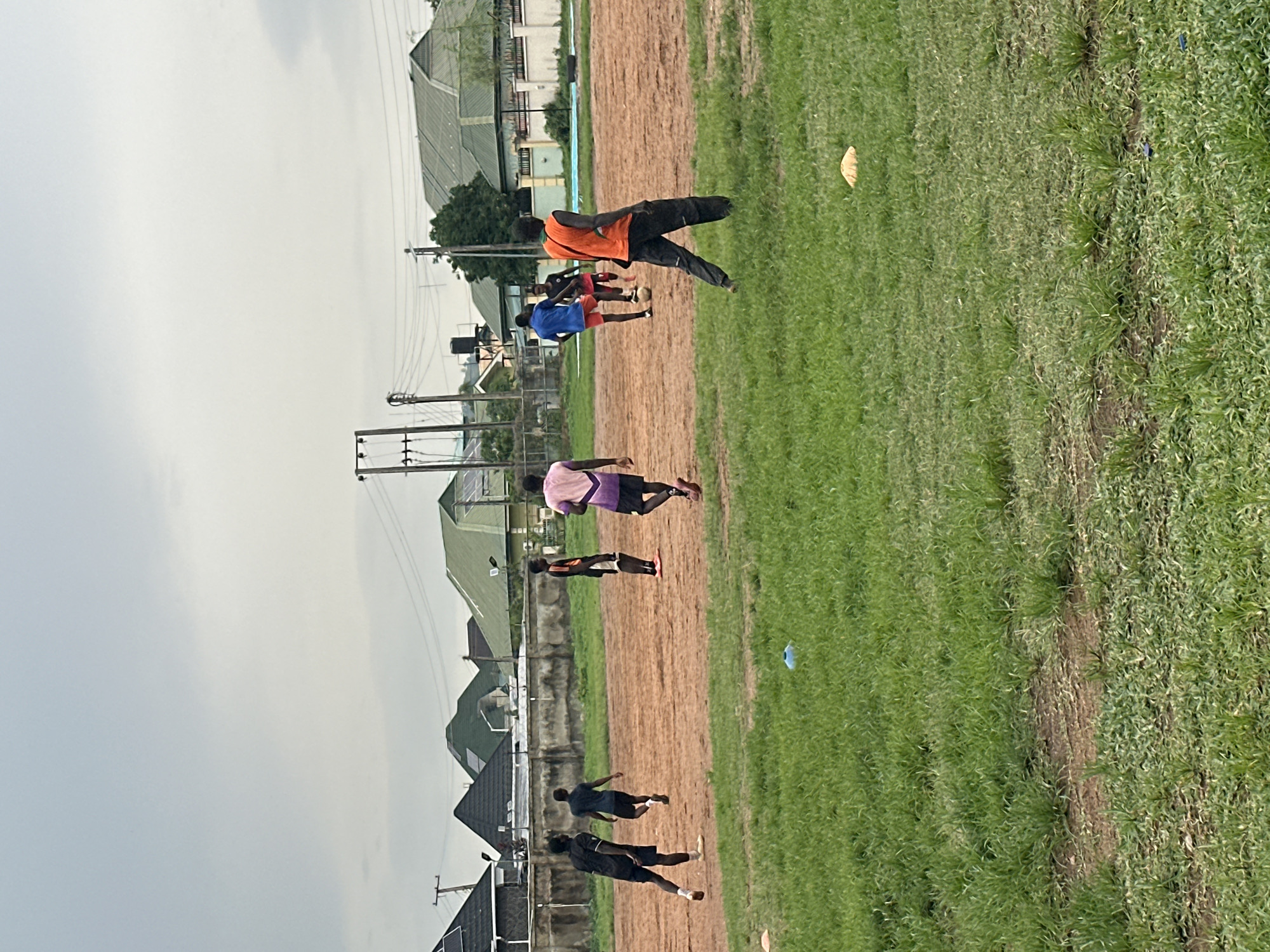 The media file shows an outdoor scene with several young men on a dirt and grass field, possibly a sports ground. The sky is overcast, suggesting a cloudy day or early morning/late afternoon. In the background, there are buildings with green roofs and what appear to be power lines and utility poles, indicative of a semi-urban or developing area.
The main subjects are the individuals, dressed in athletic wear. One person in the foreground is wearing an orange vest over dark clothing, appearing to be in motion. Other individuals are scattered across the field, some in groups and others more spread out, suggesting a training session or informal game is taking place. Their postures and the presence of a light-colored object on the grass that resembles a ball or training marker imply physical activity.
The overall setting seems to be a sports field in Abuja, Nigeria, characterized by a mix of cultivated grass, bare earth, and surrounding infrastructure. The lighting is diffused due to the cloudy weather. No text is visible in the image. The atmosphere appears casual and focused on the activity at hand.