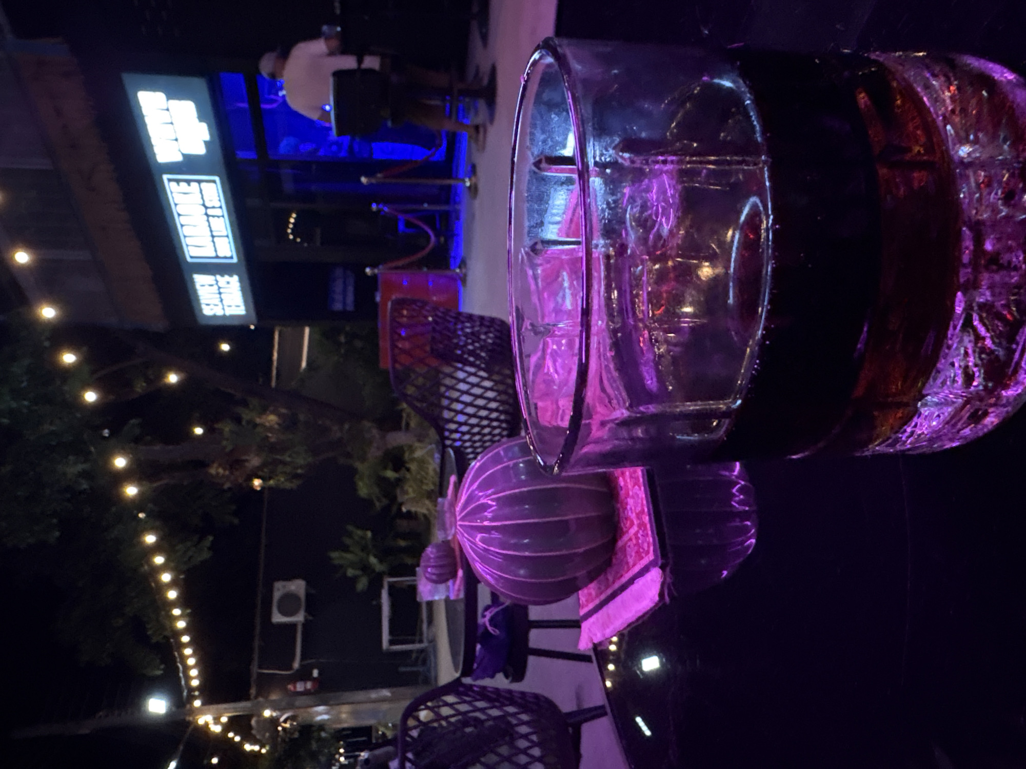 The image depicts a lit glass with liquid in front of the Seiview Terrace karaoke establishment in Baan Bang Rak, Thailand. String lights are visible in the background. In the background, a person is present behind a velvet rope barrier. A decorative sphere rests on a table in the midground. The scene is captured at night.