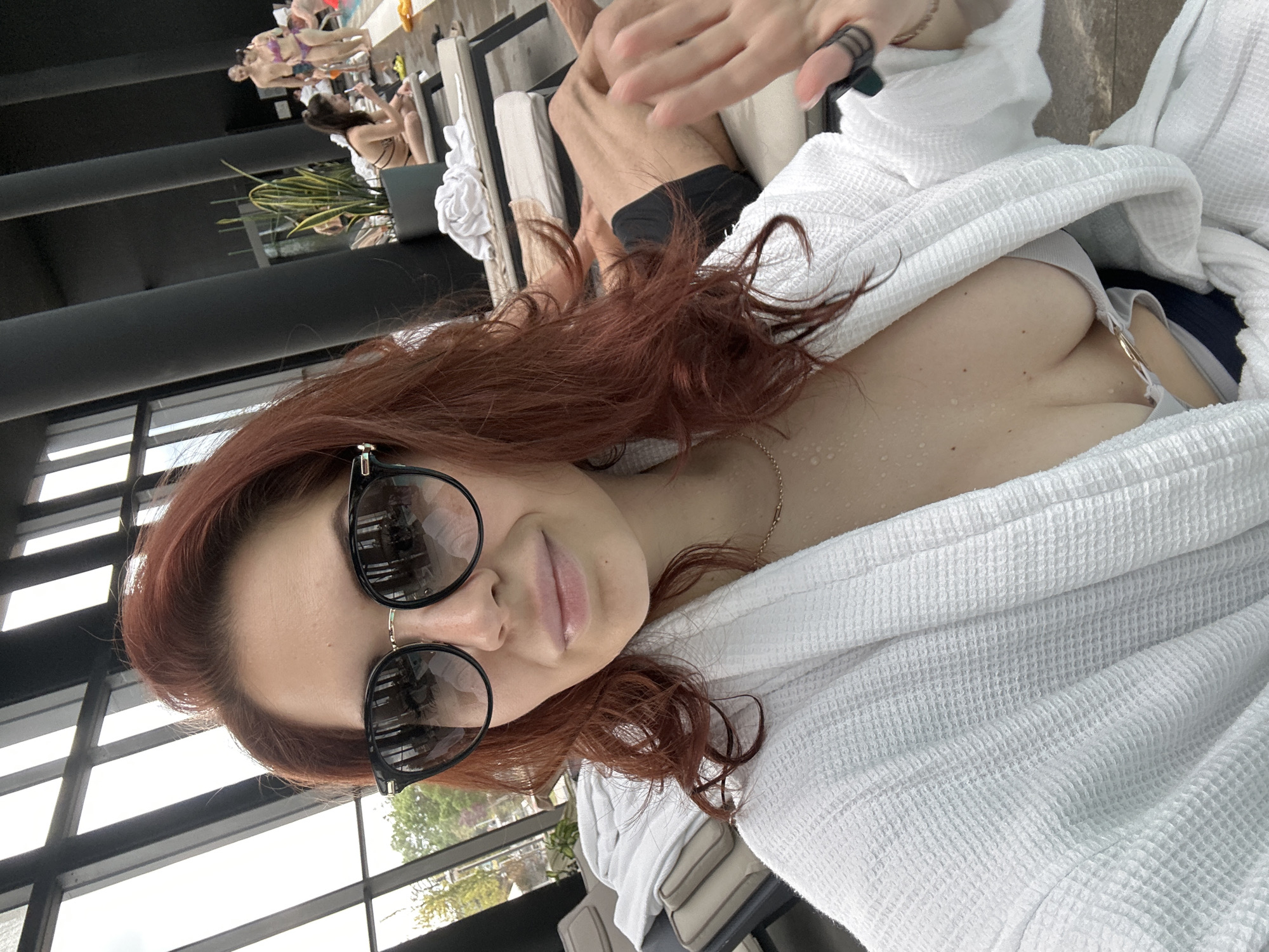 A selfie shows a red-haired woman wearing sunglasses and a white robe. Her skin appears damp. The background includes a windowed area, other people lounging by what seems to be a pool, and a black railing. A man's arm is visible in the background. Based on the tags, this image may have been taken in Kazanlak, Bulgaria.