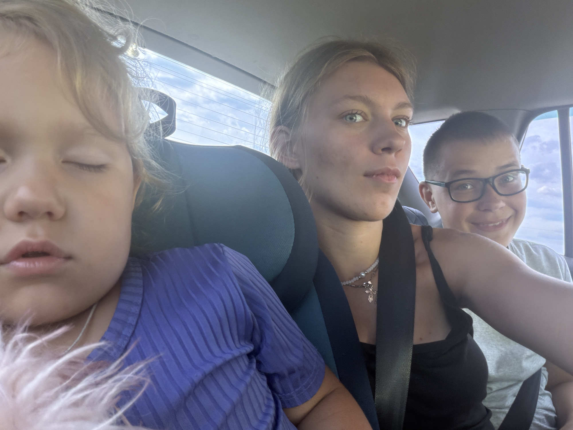 In an indoor setting, specifically within a vehicle, three individuals are visible, captured in a close-up, selfie-style photograph. On the left, a young child with eyes closed, appears to be asleep, wearing a purple, ribbed t-shirt. The middle individual, a young woman, looks directly at the camera with light blue eyes and is wearing a black tank top with a necklace featuring a cross pendant. To the right, a young boy with short hair and glasses is also looking at the camera, with a slight smile. He is wearing a light gray t-shirt. The background suggests they are traveling, with the tinted window of the vehicle and a glimpse of the sky visible. The image is well-lit, likely from natural light entering the vehicle. The context of Novoe Feizullovo, Russia, is not depicted in the visual information provided in the image.