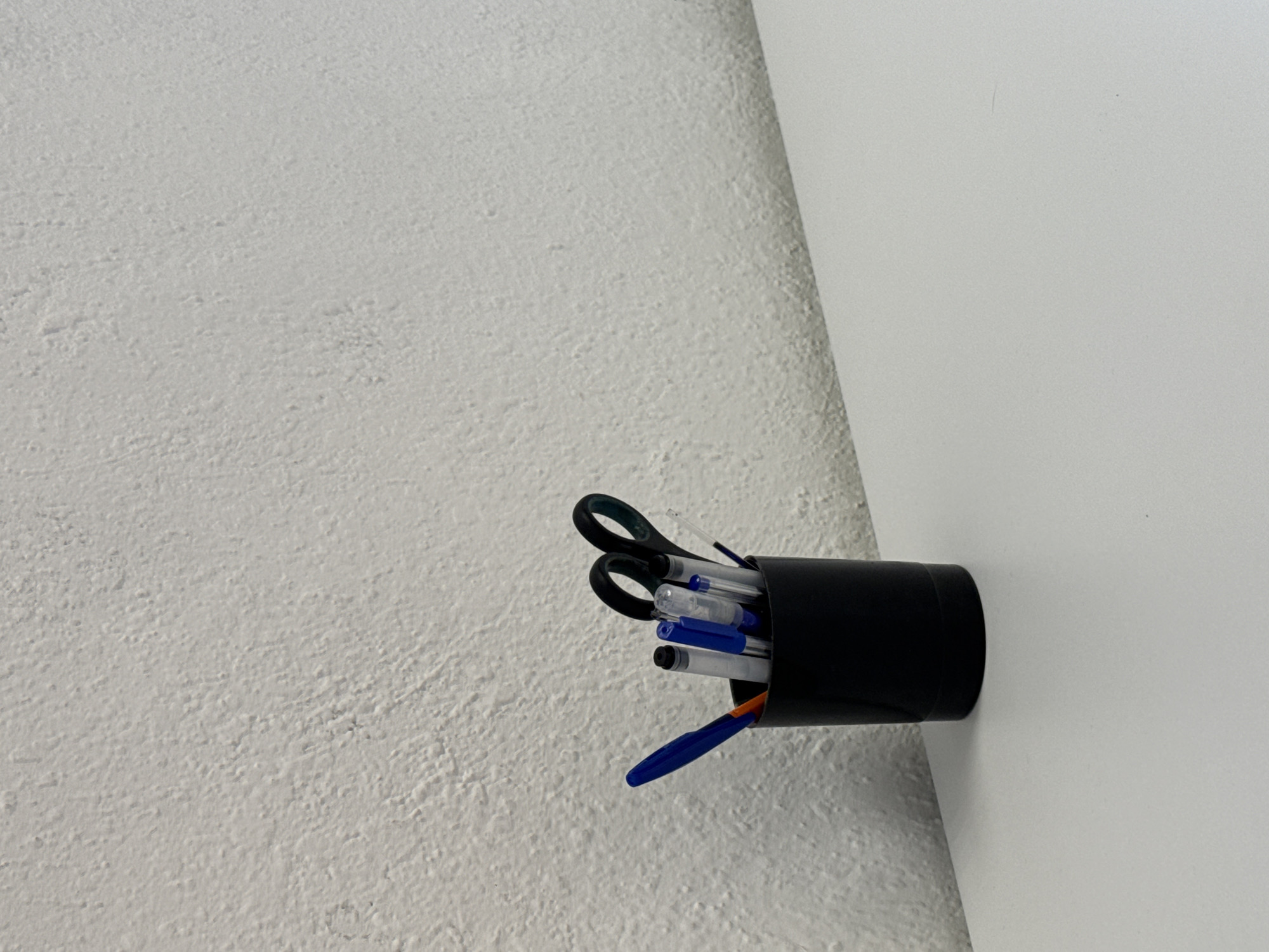 A black cylindrical container is mounted on a white wall. The container holds several pens and a pair of scissors. The wall surface is textured. The location is unknown.