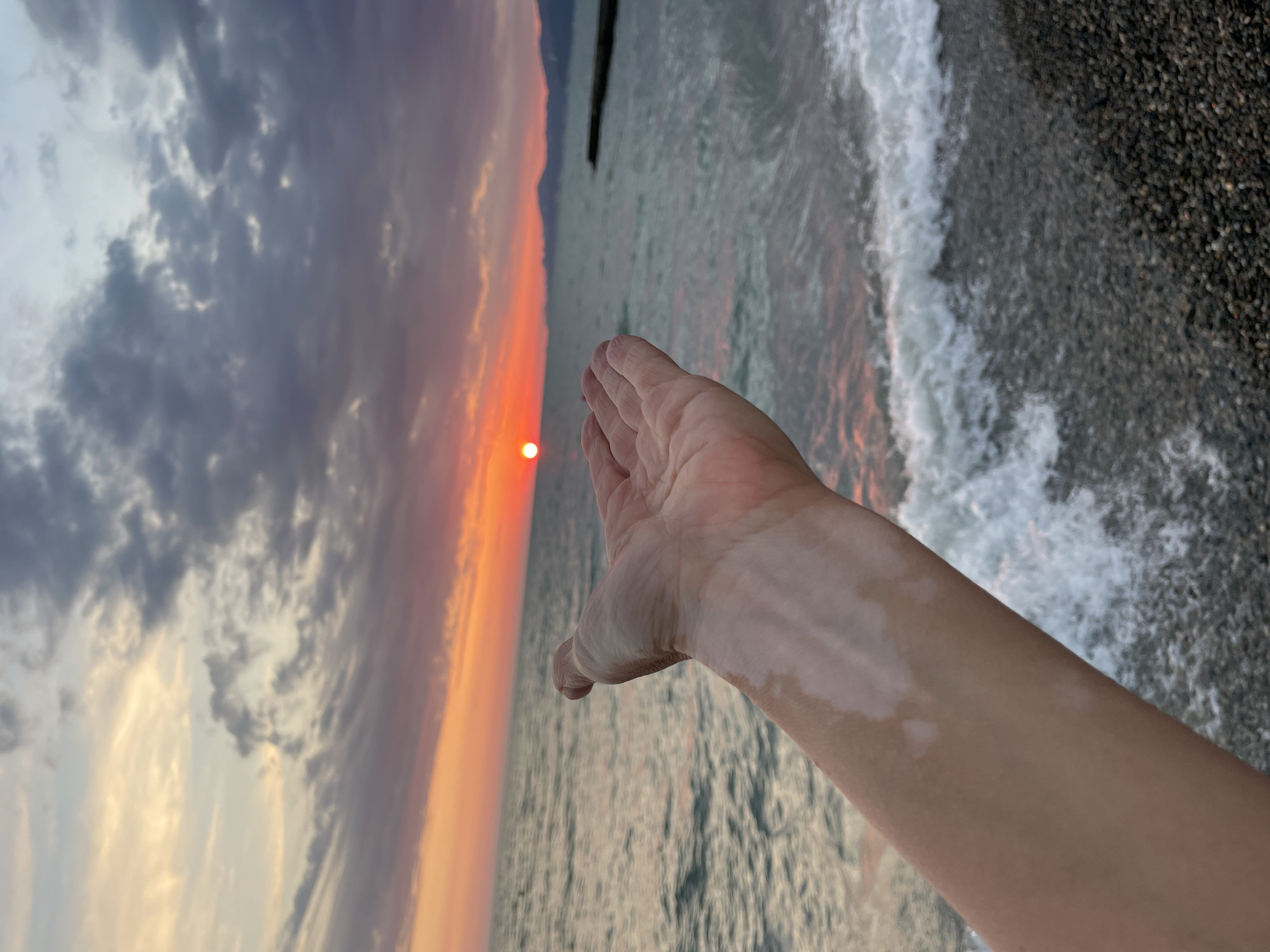 A first-person perspective shot captures an outstretched hand and forearm against a backdrop of a sunset over the ocean. The sun, a vibrant orange orb, is low on the horizon, casting a warm glow across the sky and reflecting on the water. The sky is filled with dramatic clouds, painted in hues of orange, pink, and gray. Gentle waves lap against a pebble beach in the foreground, and a dark structure, possibly a pier or breakwater, juts out into the sea in the distance. The arm in the frame has patches of lighter skin, indicative of vitiligo.