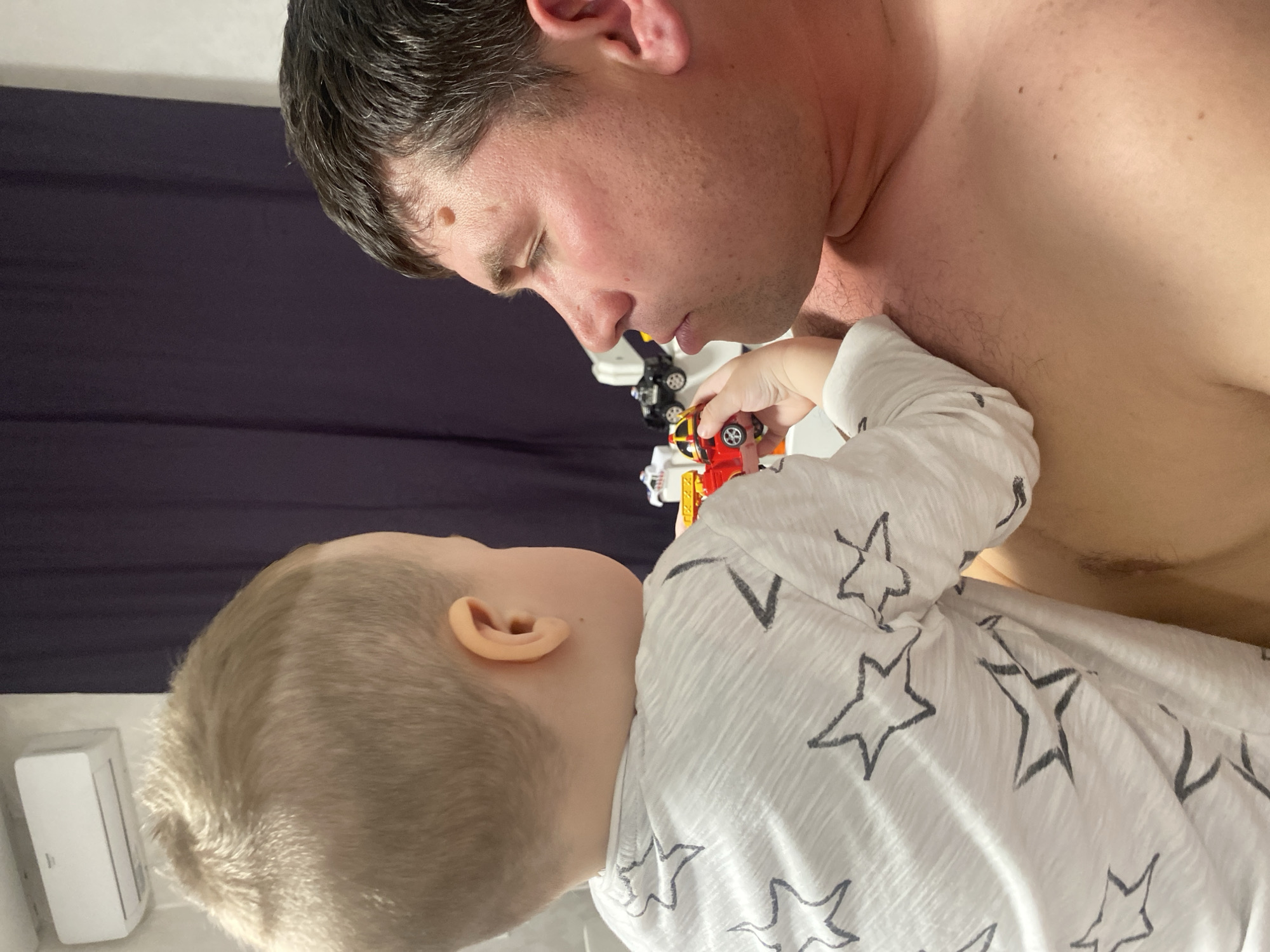 The image captures a tender, intimate moment between an adult man and a young child in an indoor setting. The photo is taken from a slightly elevated angle, looking down upon the subjects, and is oriented vertically, with the man's head on the right and the child's head on the left, but depicting them as if they are facing each other upright.
The adult, presumably the father, is on the right side of the frame. He has dark, wavy hair and fair skin, with a noticeable mole on his right temple. He is shirtless, revealing a broad chest with light hair and several moles or freckles scattered across it. His face is turned towards the child, with his eyes closed or looking downwards intently, suggesting focus or deep engagement. His lips are slightly parted.
The child, a young boy with very short, light blond hair, is on the left side of the frame. He is wearing a white long-sleeved shirt adorned with a repeating pattern of black or dark gray stars. The child's face is also turned towards the man, looking up at him with an open expression. His left ear is clearly visible.
The core of the image's activity revolves around a playful interaction. The child is holding two small toy vehicles in his right hand – one is primarily red and yellow, and the other is black. He is extending his arm, bringing these toys close to the man's chest or shoulder, as if presenting them or engaging in play. The man's posture and downward gaze indicate he is attentive to the child and the toys.
The background is simple and somewhat blurred, consisting mainly of a dark purple or navy blue fabric, possibly a curtain or bed linen. To the far left, behind the child's head, a white, rectangular object with some textured detail, possibly an air conditioning unit or a humidifier, is visible. The lighting is soft, creating a gentle and warm atmosphere.
The overall mood of the image is one of affection, connection, and parental engagement, highlighting the close bond between the two individuals as they share a quiet moment of play. There is no visible text in the image.