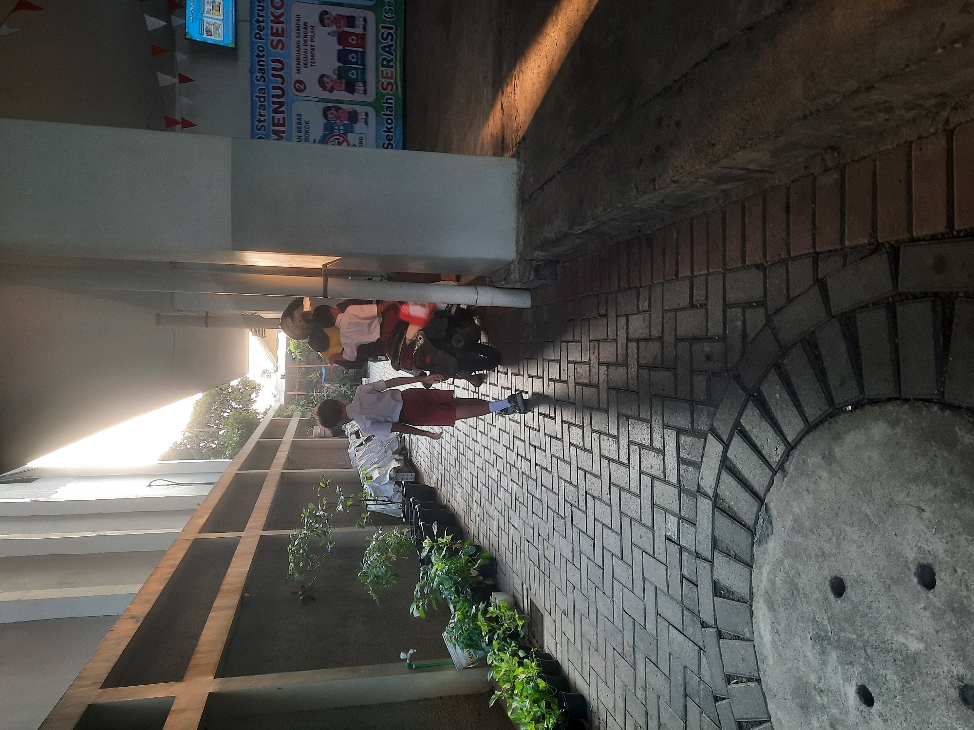 The image depicts a sunlit paved walkway lined with potted plants on the left and a building on the right. A young boy in a white shirt and red shorts, appearing to be a student, walks away from the camera down the walkway. Behind him, a few other figures are visible near a parked scooter. The building on the right features a poster with Indonesian text that includes the words "Sekolah SERASI". Red and white triangular flags are strung along the top of the building. The environment suggests an outdoor school or institutional setting. The lighting indicates it is daytime, with sunlight casting long shadows across the pavement.