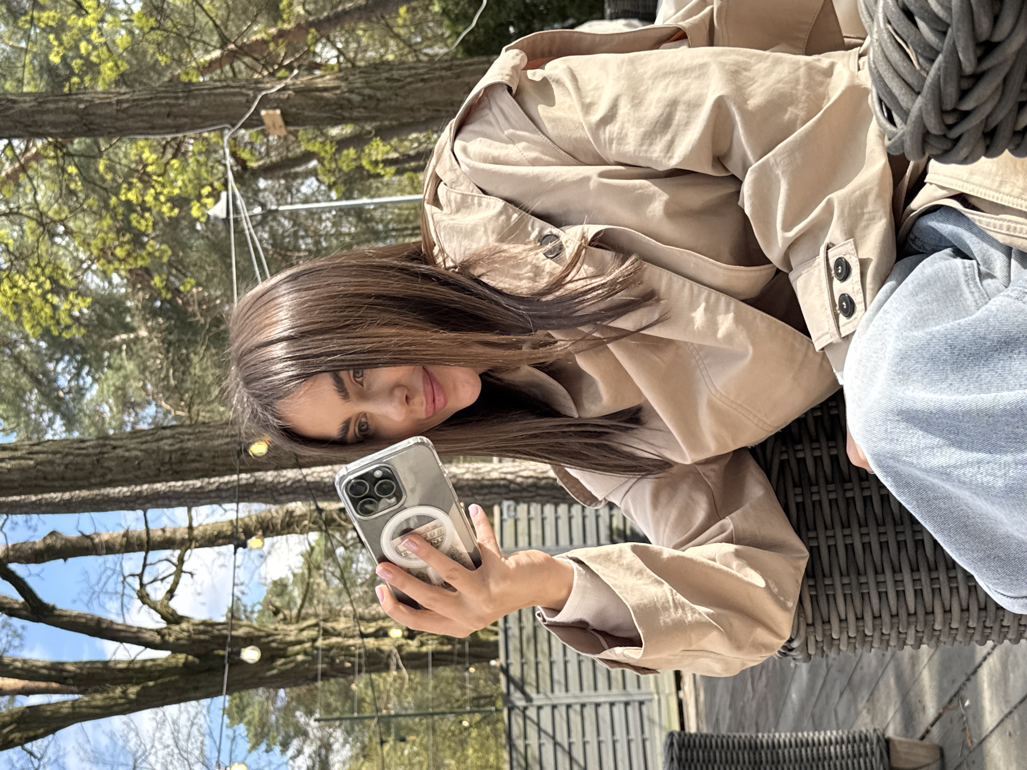 A woman with long, brown hair and a tan trench coat is sitting at an outdoor table, holding a smartphone. The phone has a clear case with a circular MagSafe ring and what appears to be a picture or design visible beneath the ring. The woman is looking at the phone screen. Behind her are trees with bare branches and string lights. A blue denim garment is partially visible at the edge of the table. The table itself is made of dark woven material. This scene is in an outdoor setting with trees and string lights. The location is Konstancin-Jeziorna, Poland.