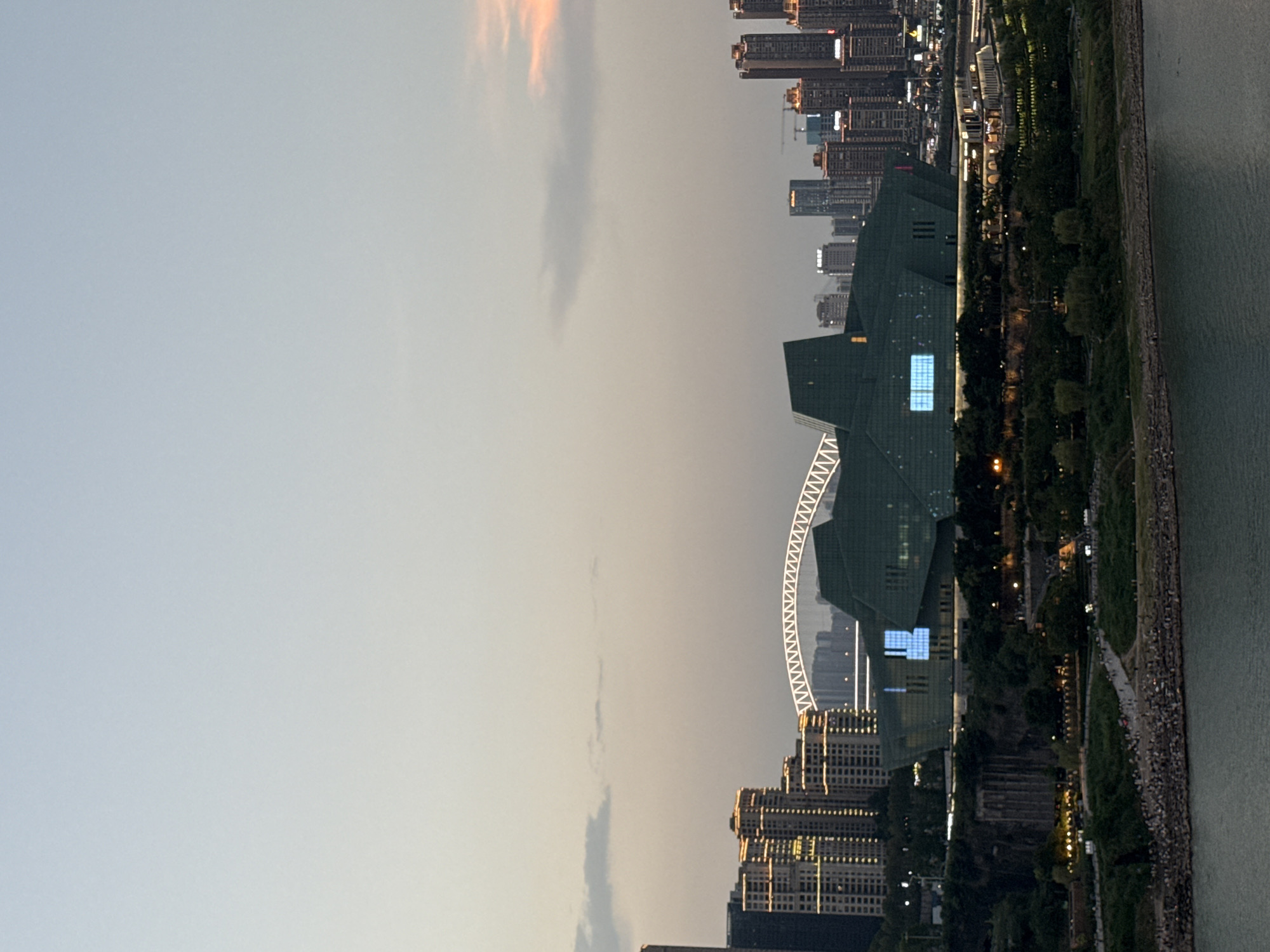 An elevated, wide shot captures a city skyline at dusk, likely in Yuzhong District, China, with a river in the foreground. The sky is a pale gradient of blue and gray, with a hint of peach on the right horizon. A large, angular, dark green building with illuminated blue windows dominates the midground, set against a backdrop of numerous high-rise buildings, many of which are lit with warm yellow lights. A white arched bridge with a zigzag pattern is visible behind the green building and among the skyscrapers. To the left of the green building are a cluster of taller, lighter-colored buildings also illuminated. The immediate foreground of the midground shows a rocky shoreline with many small figures of people scattered across it. Beyond the shoreline is a lush green area with trees, pathways, and more illuminated structures, suggesting a park or public space. The river in the foreground is a calm, dark blue-green. The overall scene depicts an urban environment transitioning from day to night, with architectural landmarks and a significant population present.