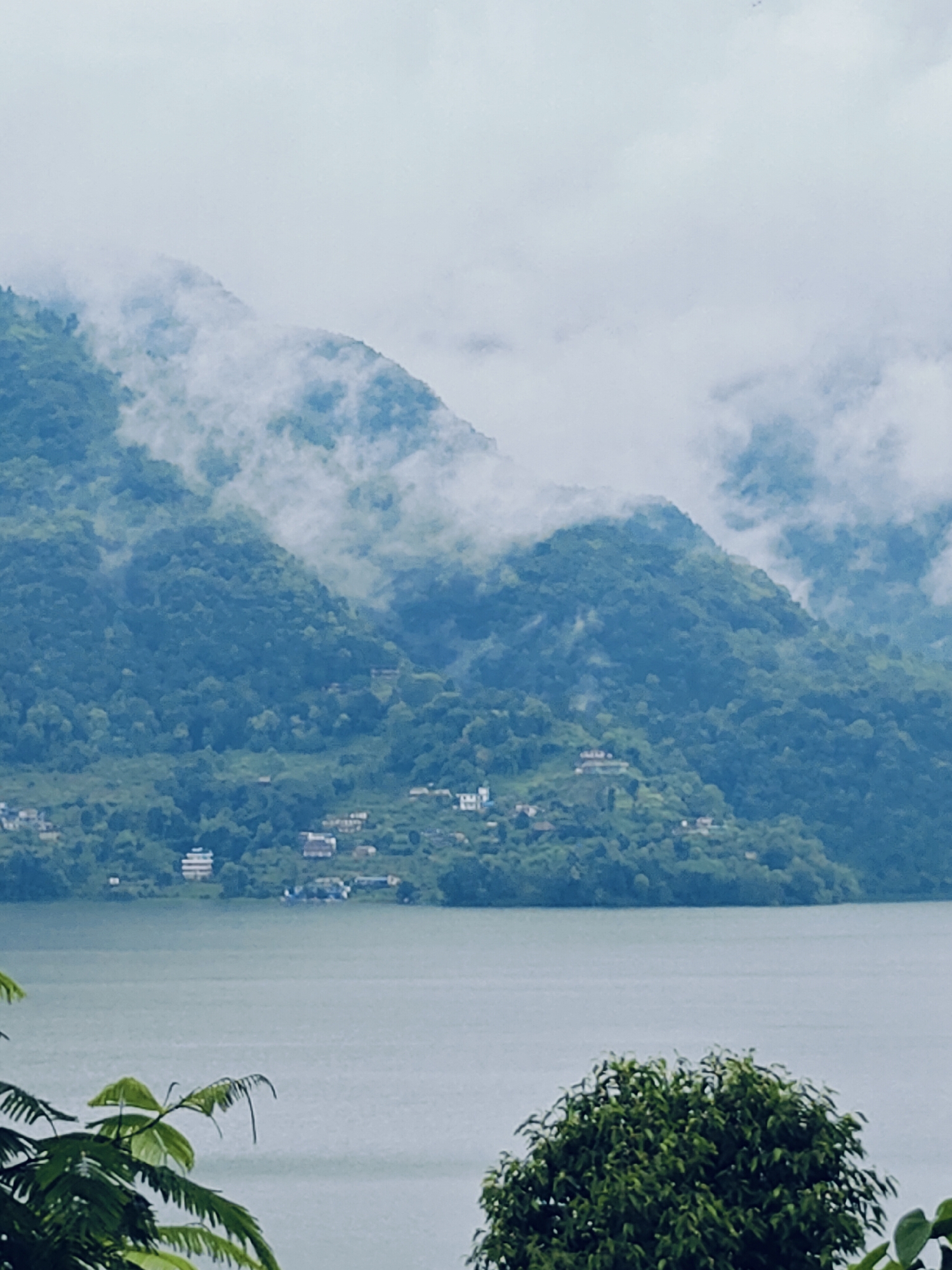 A serene, misty landscape captures the tranquility of Pokhara, Nepal. The image features a wide, calm body of water in the foreground, reflecting the overcast sky. Lush, tree-covered mountains rise dramatically behind the water, their slopes partially obscured by swirling white clouds. Small clusters of buildings dot the hillsides, hinting at a settlement nestled within the natural beauty. The overall impression is one of peaceful isolation and the majestic power of nature.