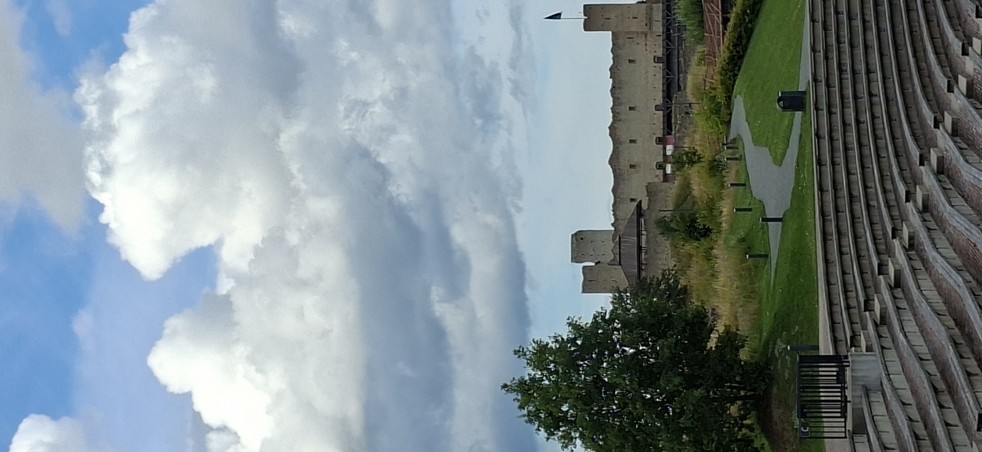 The image depicts a view of Rakvere Castle in Rakvere, Estonia. The castle, with its stone walls and turrets, stands against a partly cloudy sky with large, white clouds dominating the upper left portion of the frame. The scene is oriented vertically, with the castle appearing on the right side of the image.
In the foreground, to the left of the castle, is a lush green tree with dark foliage. To the right of the castle, a pathway winds through a grassy area, leading towards the castle. Beyond the grass, a set of tiered seating, possibly an amphitheater or grandstand, made of brick and stone, fills the right side of the image. The seating appears to be constructed in a curved, layered fashion.
The lighting suggests it is daytime, possibly late morning or afternoon, with soft light illuminating the scene. The weather appears to be pleasant, with a mix of sun and clouds. There are no people visible in the image, and the primary subjects are the historical castle, the surrounding landscape, and the architectural elements of the seating area. The overall impression is one of a historical site and public space bathed in natural light.