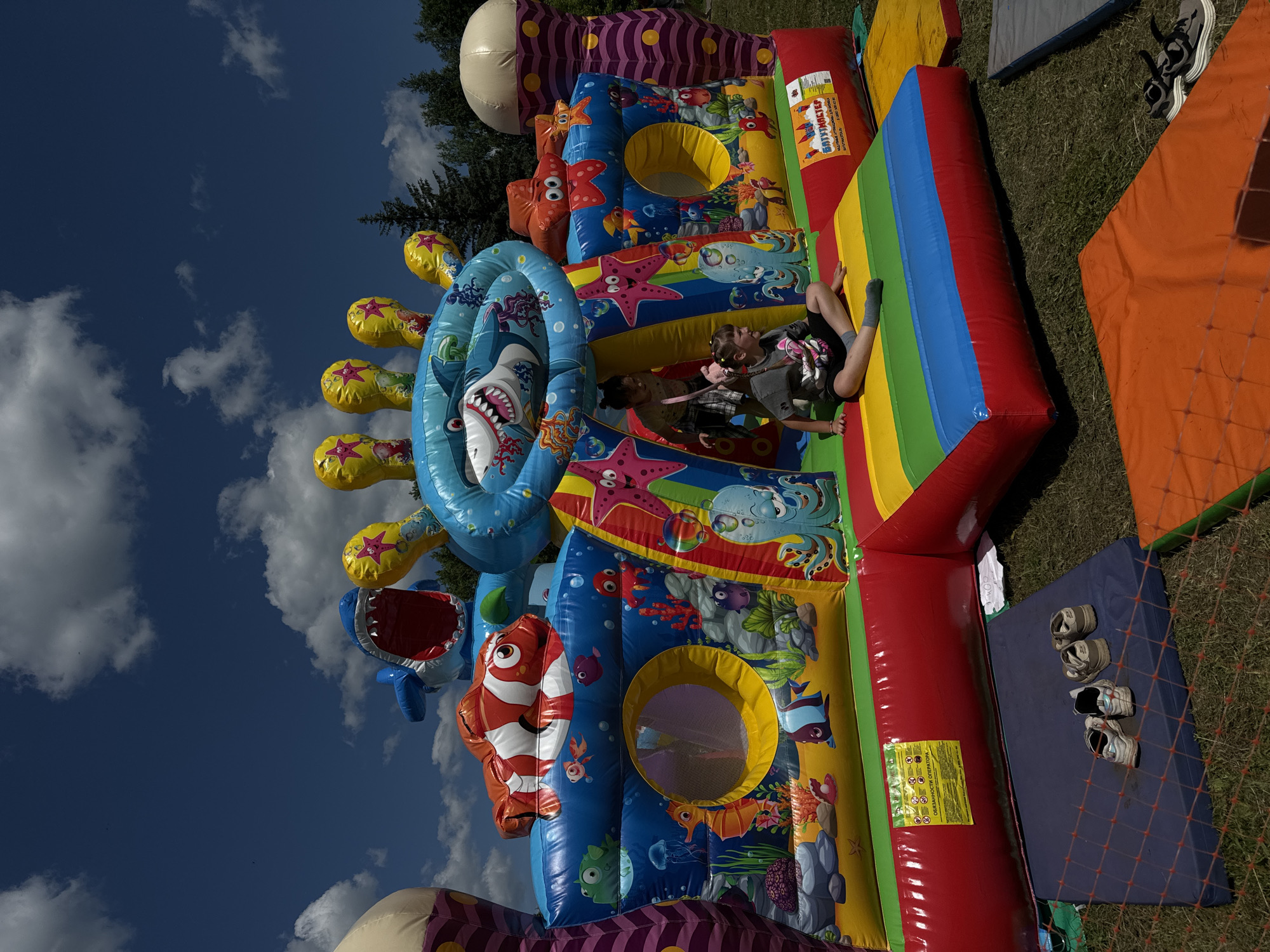 The image shows a colorful, inflatable water-themed amusement structure in an outdoor setting. The structure is decorated with various sea creatures, including sharks, starfish, and fish. Two children are visible on top of the structure, appearing to be playing or resting. The scene is set on a sunny day with a clear blue sky and some clouds. The ground appears to be covered in grass, and there are some additional inflatable pieces and mats nearby. Several pairs of shoes are neatly placed on a blue mat on the grass, suggesting the children removed them before entering the inflatable. A sign with text is visible on the side of the inflatable. The overall impression is one of a fun outdoor activity for children. The location context provided is Ponasirevo, Russia.