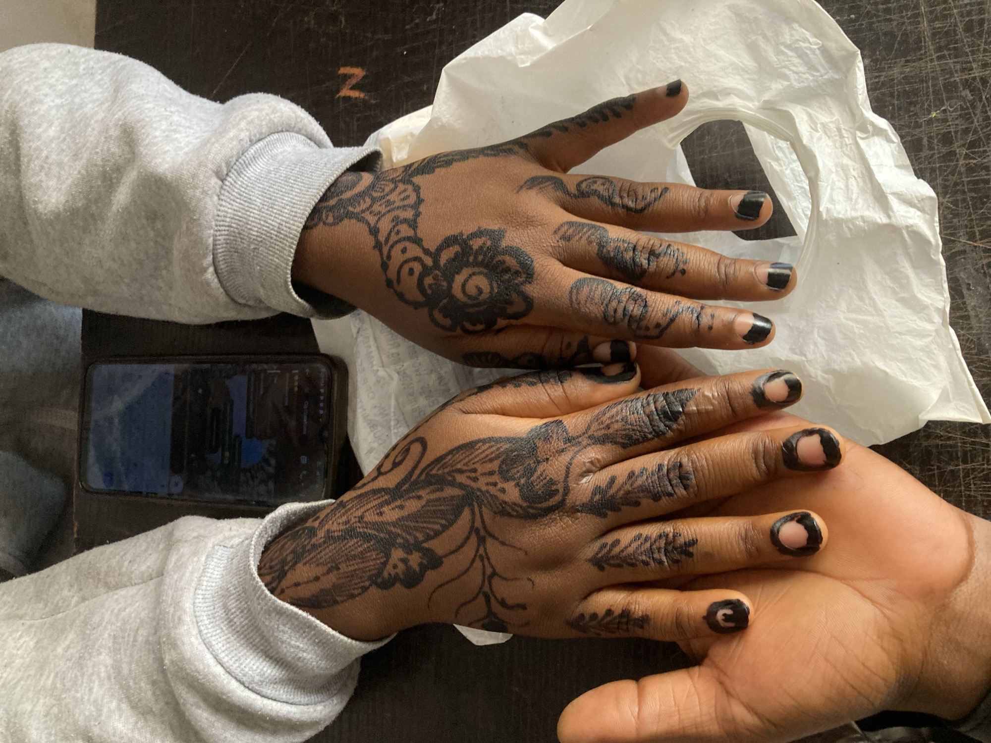 The image displays a close-up, overhead view of two hands adorned with intricate dark henna (mehndi) designs. The hands belong to an individual with a medium-dark skin tone, likely in Jos, Nigeria, where henna is a common form of adornment.
The left hand, positioned further back, features a prominent floral motif with swirling petals and surrounding decorative elements on the back of the hand. The design extends up the wrist and onto the fingers, where lines and patterns cover the knuckles and phalanges. The fingertips are also entirely covered in the dark henna, resembling a solid nail polish. The hand emerges from the cuff of a light grey, ribbed, long-sleeved sweatshirt.
The right hand, closer to the foreground and slightly overlapping the left, showcases a different, more flowing design on its back, characterized by leaf-like patterns and vine-like tendrils. Similar to the left hand, the fingers are decorated, and the fingertips are fully darkened with henna. The palm of the right hand is visible, showing clear skin without henna.
Between the hands, towards the upper right, is a crumpled piece of white material, possibly a plastic bag or tissue. On the left side, beneath the left wrist, a smartphone is partially visible, its screen illuminated and displaying what appears to be a messaging interface with blue and white elements. The background is a dark, textured surface, possibly a wooden table. A faint orange letter "Z" is discernible on this surface above the left hand. The overall impression is one of fresh, richly applied henna, suggesting a recent application or celebration.