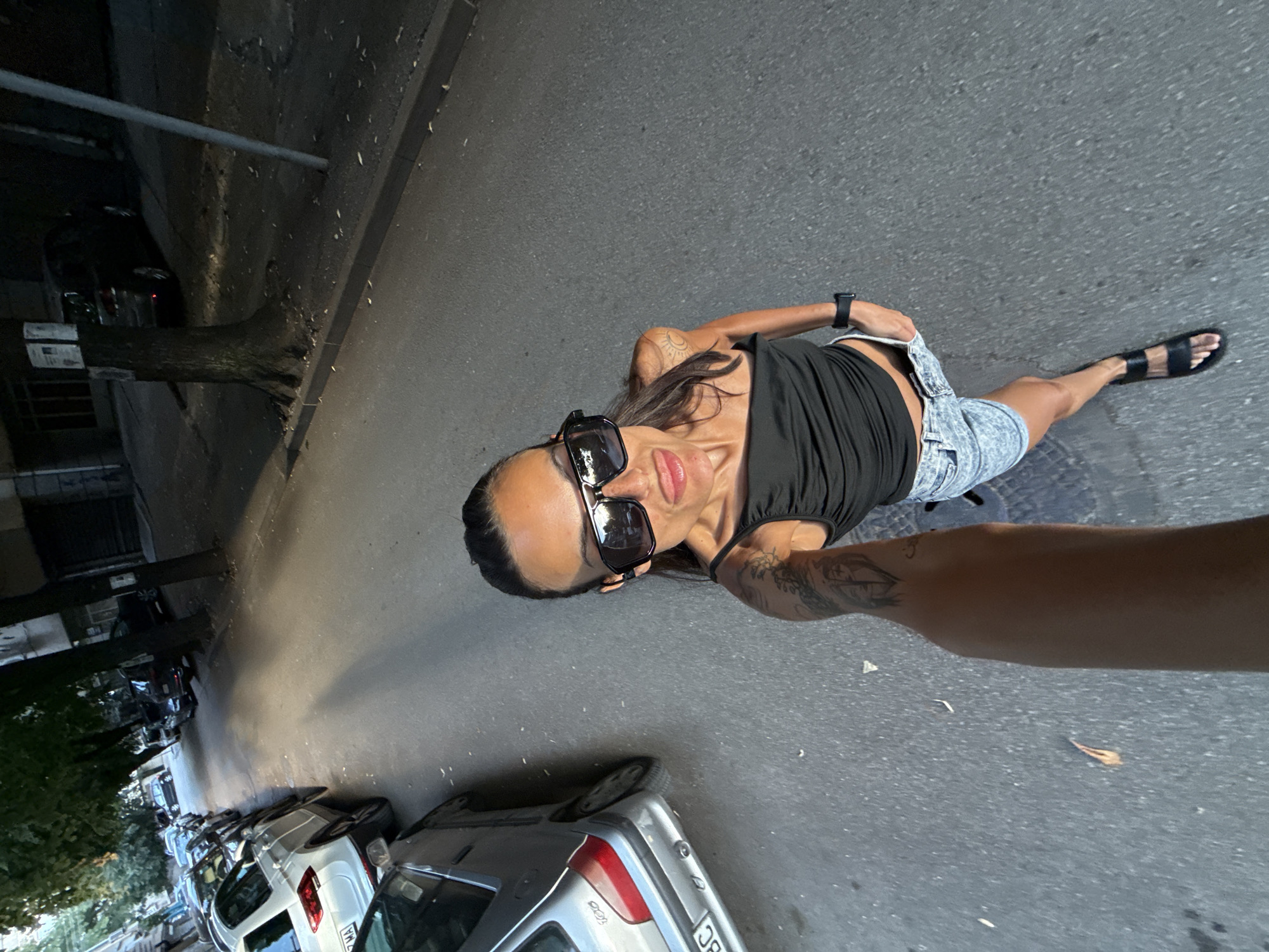 A woman wearing large sunglasses, a black tank top, and denim shorts is taking a selfie on a street in Pleven, Bulgaria. She is sticking her tongue out playfully. Cars are parked along the street, and trees line the sidewalk. The sun is shining, casting shadows on the ground. A dark pole is visible to the right of the frame.