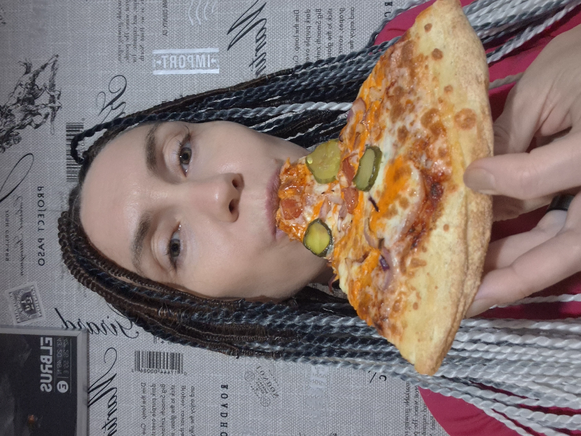 The image features a person with braided hair holding a slice of pizza to their mouth. The person's face is partially visible, and they appear to be in the process of taking a bite. The pizza has visible toppings, including cheese, tomato sauce, and pickles. The background is a patterned fabric, possibly a tablecloth or a wall covering, with text and graphic elements that are difficult to decipher clearly due to the image's orientation and focus. Some visible text includes "TAORM" and what appears to be Cyrillic script. The lighting suggests an indoor setting, and there are no strong indications of the time of day or weather. The overall impression is casual and relates to food consumption.