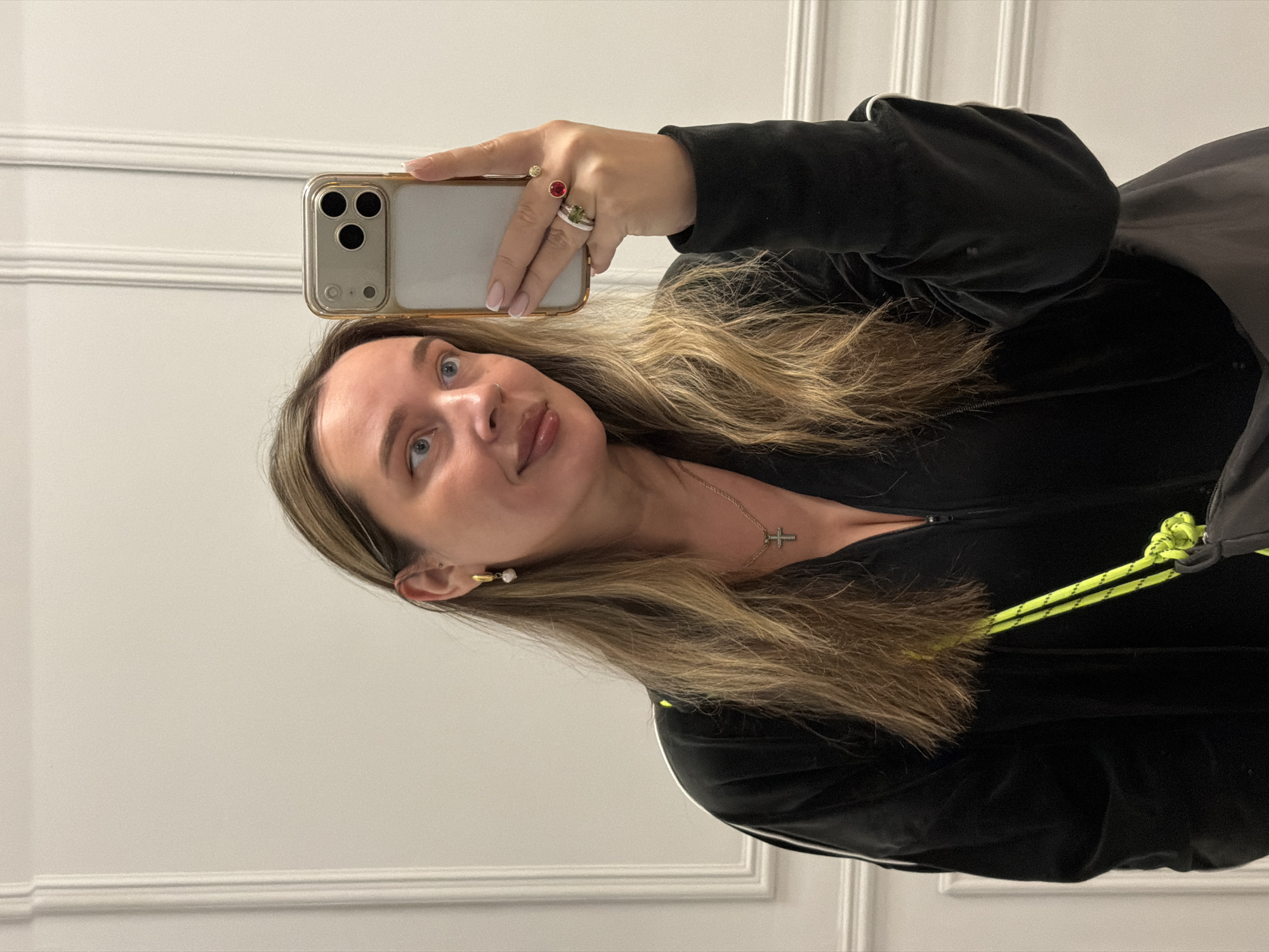 A woman takes a selfie indoors. She is wearing a black jacket with lime green drawstrings, a gold cross necklace, and earrings. She has blonde hair. She is holding a gold iPhone with three cameras in her right hand, on which she is wearing multiple rings. The background consists of a white wall with panel molding. No information suggests the location is Moscow, Russia.