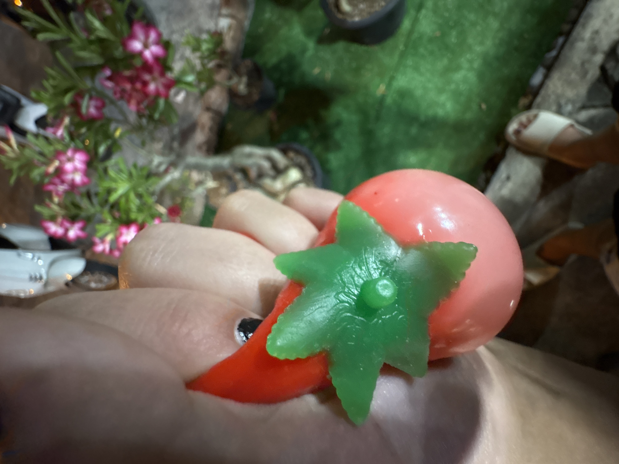 A hand squeezes a pink toy tomato with a green leaf top, as a white scooter and flowering plants are visible in the background. The toy is held by the thumb and forefinger, with a portion of the red stem of the toy also visible. The background shows a blurred image of a white scooter, a potted plant with pink flowers, and a patch of green artificial turf. A person's feet wearing white sandals are also visible at the bottom right of the image. The scene appears to be outdoors at night.