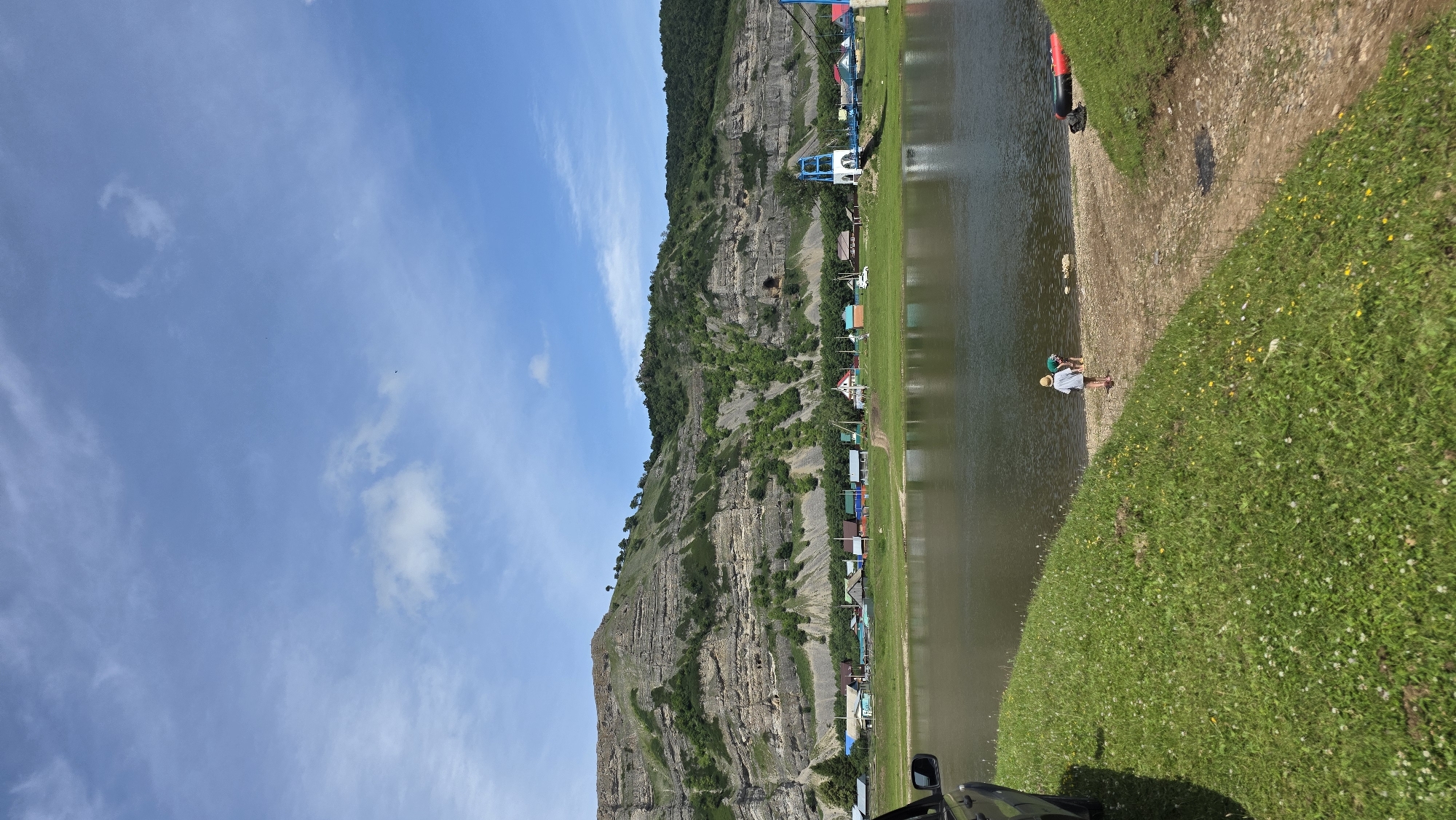 The image depicts a scenic riverside view in the village of Tash-Asty, Russia. A calm body of water, likely a river, dominates the foreground, reflecting the blue sky and clouds. On the far bank, a small village is nestled against a large, layered rock formation and lush green hills. The village consists of several colorful houses with red, blue, and green roofs. A white suspension bridge with blue supports spans across the river near the village. In the foreground, a grassy bank slopes down to the water. Two children, one wearing a hat and the other in a green shirt, are near the water's edge, with a raft lying on the bank to the right. A dark-colored vehicle is partially visible on the left side of the frame. The scene suggests a peaceful, rural setting on a bright, sunny day.