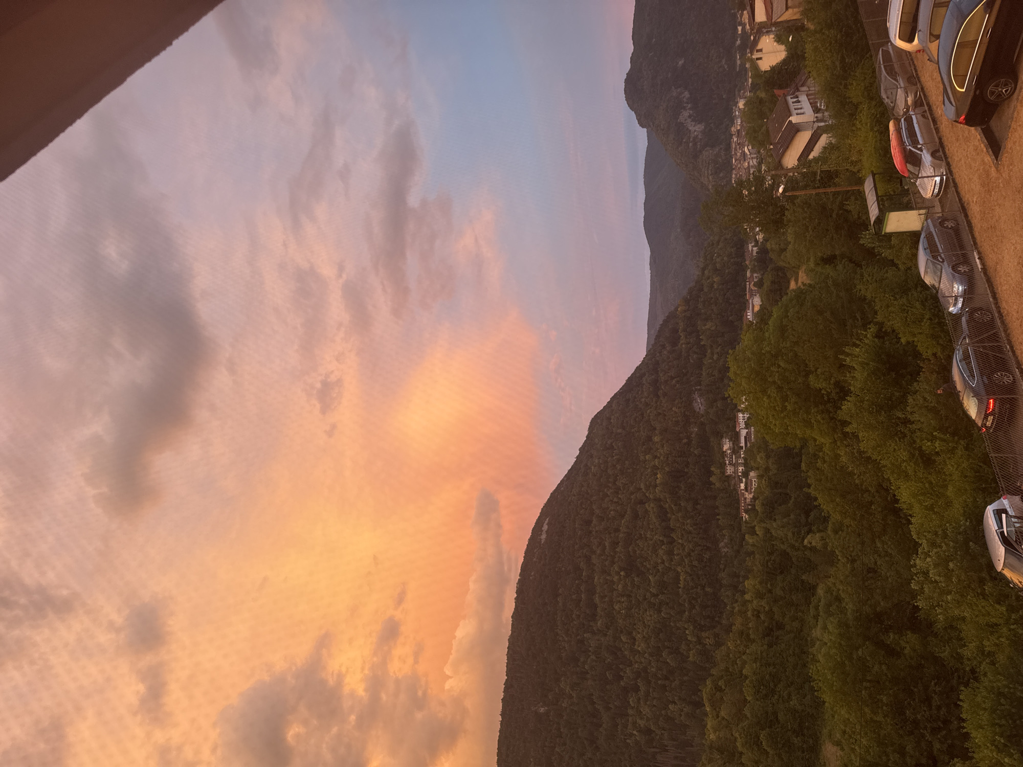 The image depicts a sunset over the city of Saint-Claude, France. The sky is illuminated with vibrant hues of orange, pink, and blue, with scattered clouds. In the foreground, a row of parked cars is visible, with a fence separating them from a dirt area. Beyond the cars, a lush green forest covers the slopes of a mountain, with a small town nestled amongst the trees. The town features buildings with red-tiled roofs. In the distance, more rolling hills and mountains are silhouetted against the colorful sky. A single person is standing near a black car on the right side of the frame. The overall scene captures the beauty of a rural French landscape at dusk.