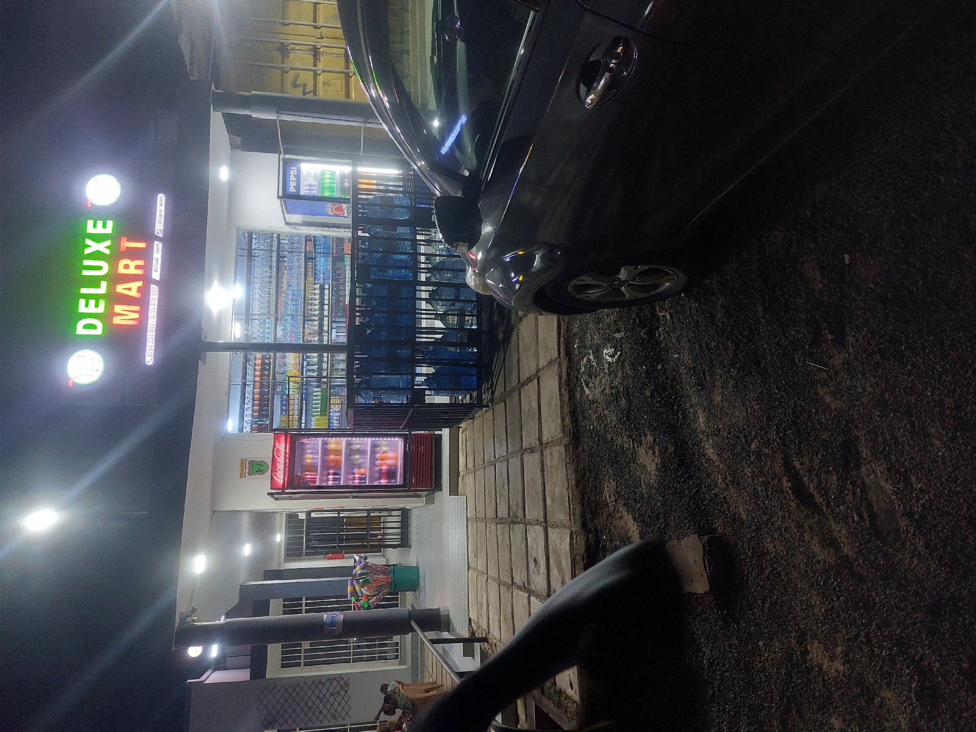 The image shows the exterior of a "Deluxe Mart" convenience store, likely at night, based on the lighting conditions. A dark-colored car is parked closely on the right. Several rows of shelved drinks are visible inside the store, as well as a red refrigerator at the back. There is an awning with a Pepsi advertisement. The store front has a bright green "Deluxe Mart" sign and phone number in the front, with another sign parallel to the store front displaying the store information. Some people are visible sitting under an awning. The location is likely Dar es-Salaam, Tanzania, based on the store phone number on the sign.