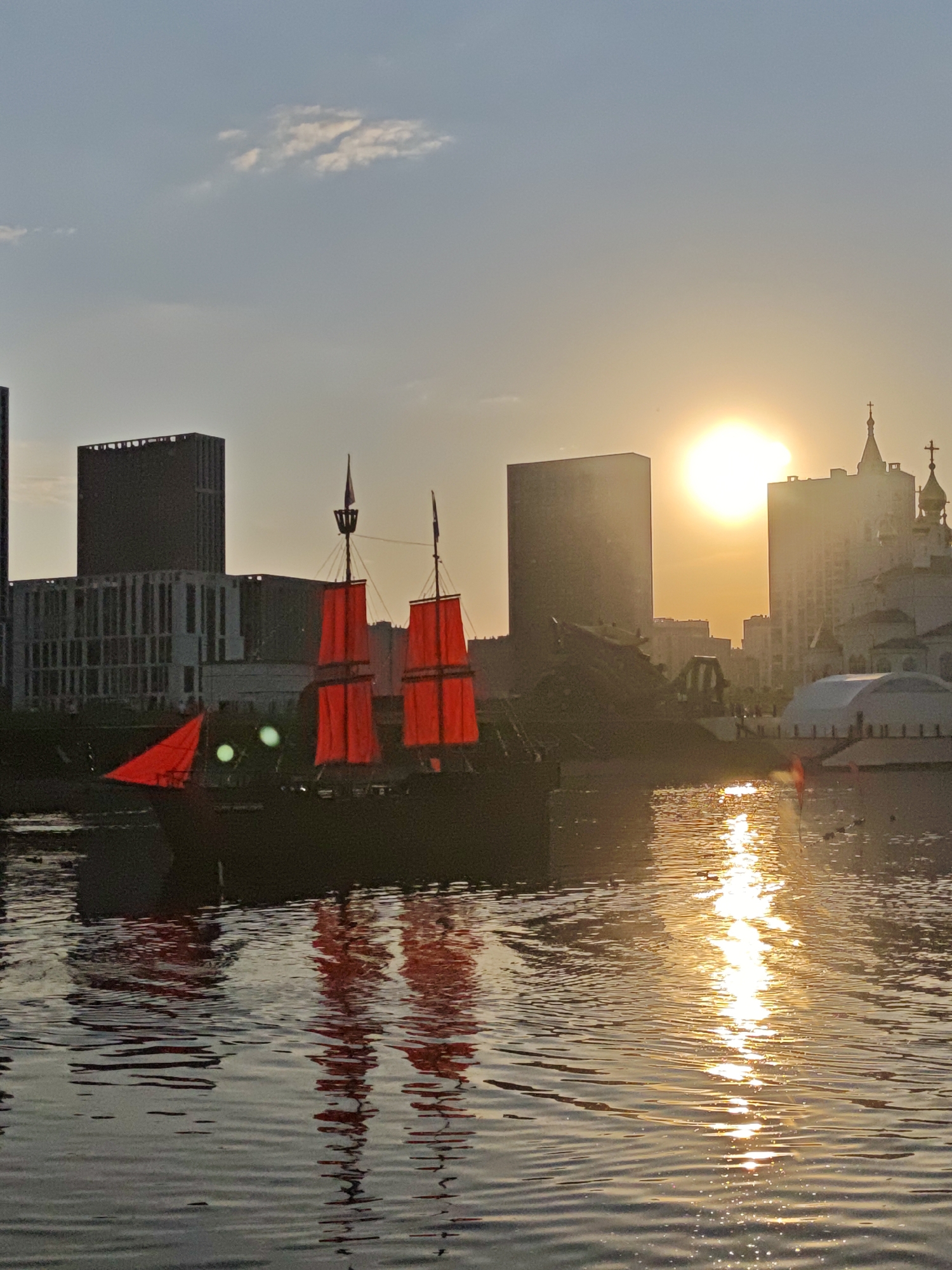 Captured in Yekaterinburg, Russia, the image presents a body of water reflecting a setting sun. A ship with red sails is the focal point, contrasted against modern architecture and a church in the background. The sunlight reflects intensely on the water, creating a bright, shimmering effect. The sky is slightly overcast.