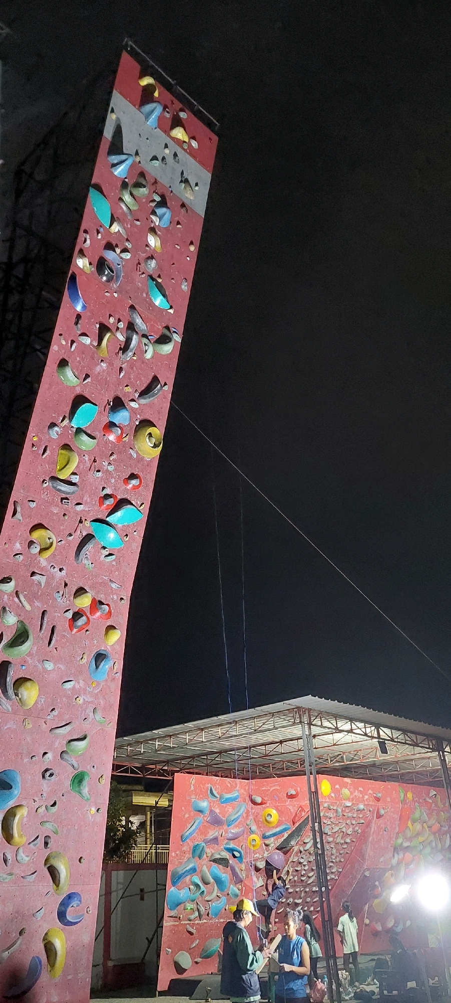 An outdoor climbing facility is illuminated at night. A tall, pink climbing wall with various colorful holds stands at an angle on the left. To the right, a wider, red climbing wall is visible under a metal-roofed shelter. Several people are present: one person is climbing the red wall, while others are standing near the base of the walls, some appearing to be instructors or observers. The sky is dark, indicating it is nighttime.
