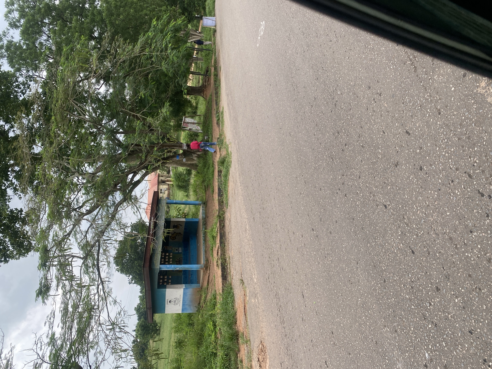 A medium shot captures a roadside bus stop on an overcast day in Ojere, Nigeria. The bus stop is a simple, open-air structure with a blue and white painted concrete building. It features a blue-tiled bench and an opening on the side facing the road. A white and blue emblem is visible on the left side of the bus stop, and a mural depicting Africa is painted on the right. The bus stop is surrounded by green grass and trees. In the foreground, a paved road stretches across the bottom of the frame. A person wearing a pink shirt and blue jeans walks away from the bus stop towards the right side of the frame. Further in the background, another person wearing a blue shirt and dark pants stands near a white sign. To the right of the bus stop, a building with a red roof is partially visible. The overall impression is of a quiet, rural bus stop.