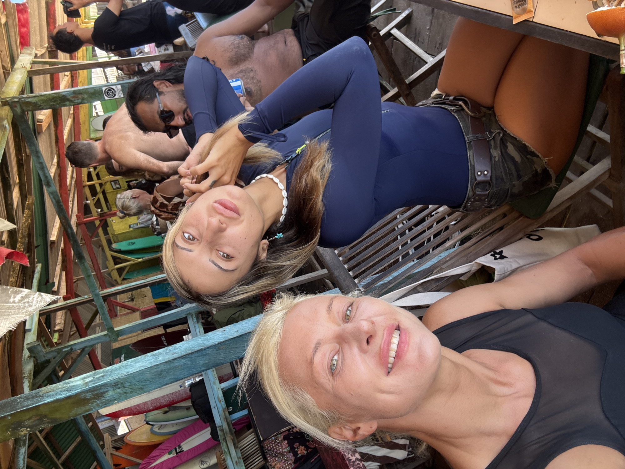 The image captures a selfie taken by a woman with short blonde hair wearing a black top. She is smiling at the camera. In the background, another woman is visible, wearing a navy blue wetsuit, a shell necklace, and camo shorts with a brown belt. Several other people are in the background, some shirtless and some wearing dark clothing. The setting appears to be an outdoor location, possibly a beach bar or restaurant in Unknown. Surfboards are visible amongst the wooden framework, suggesting a seaside environment.