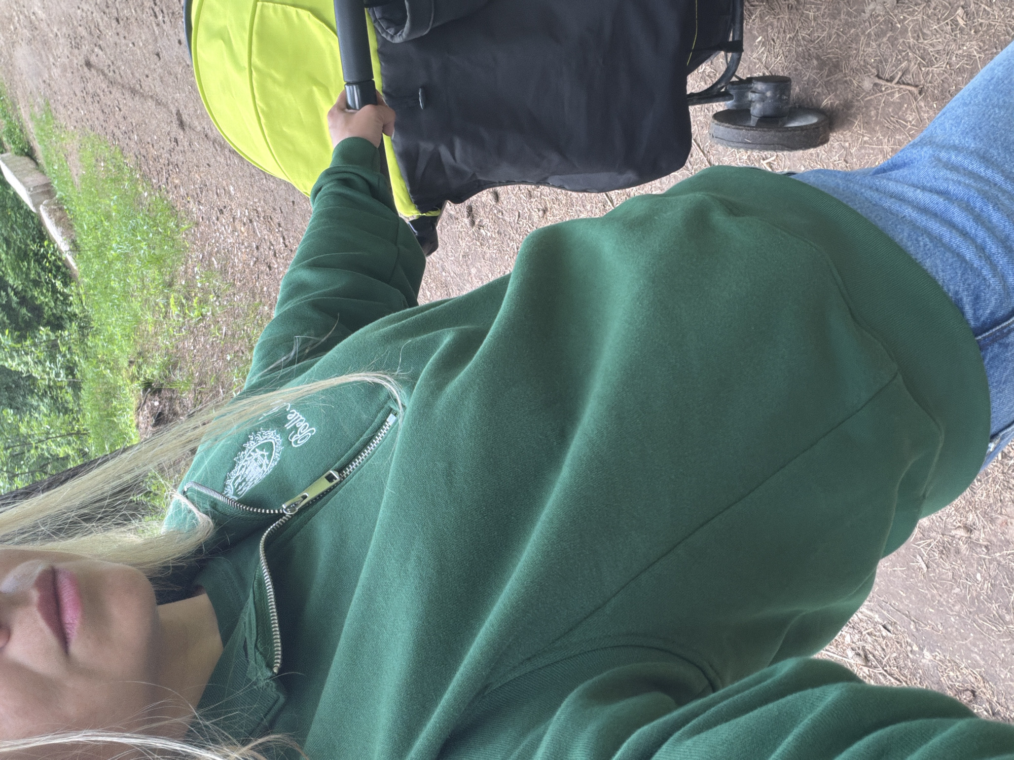 The image is a first-person perspective shot of a person pushing a baby stroller on a dirt path. The person is wearing a green zip-up hoodie and blue jeans. The stroller has a neon green seat and black storage. The path is lined with grass and trees. The location is not identifiable as Votkinsk, Russia based on visual evidence.