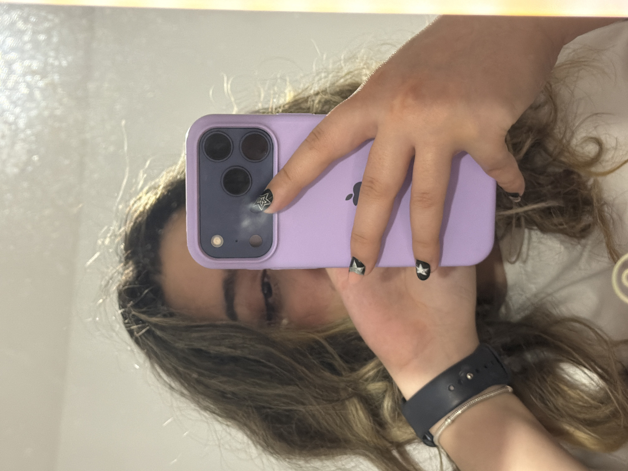 A person is captured taking a mirror selfie, partially obscuring their face with a smartphone. The subject's long, wavy brown hair with lighter highlights frames the visible portion of their face, which shows their eyes reflected in the mirror. Both hands are holding a light purple-cased iPhone, identifiable by the distinct Apple logo on its back. The phone's camera module, featuring three lenses and a flash, is clearly visible. The person's fingernails are painted black and adorned with prominent white or silver star designs. On their right wrist, a dark smartwatch band, likely an Apple Watch, is worn alongside a thin silver bracelet. The background reflected in the mirror appears to be a plain, light-colored indoor setting, possibly a wall or ceiling, with some subtle textural details. A bright light source is visible at the top edge of the reflection. The scene is located in Plovdiv, Bulgaria.