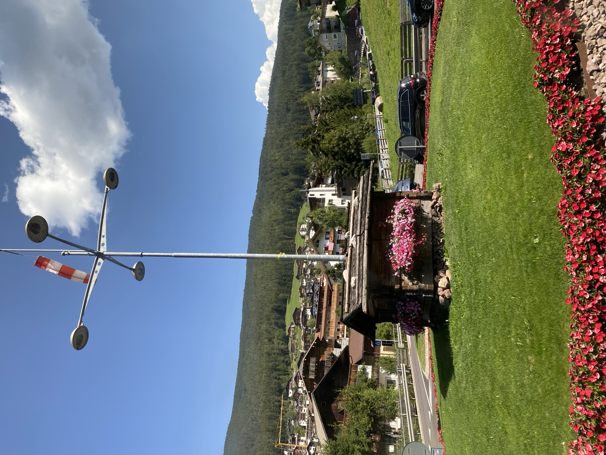 A bright, sunny day in Urtijëi - St. Ulrich in Gröden - Ortisei, Italy features a picturesque scene. A red and white striped windsock is prominently displayed on a multi-armed lamppost. Below the lamppost, a small, rustic wooden structure, possibly a decorative kiosk or information booth, is adorned with abundant pink flowers cascading from planters. The structure sits on a vibrant green lawn, bordered by a bed of bright red flowers. In the background, a charming village with traditional wooden-roofed houses climbs the side of a heavily forested mountain. A road with several cars parked and driving is visible, along with a construction crane in the distance. The sky is a clear blue with a few fluffy white clouds.