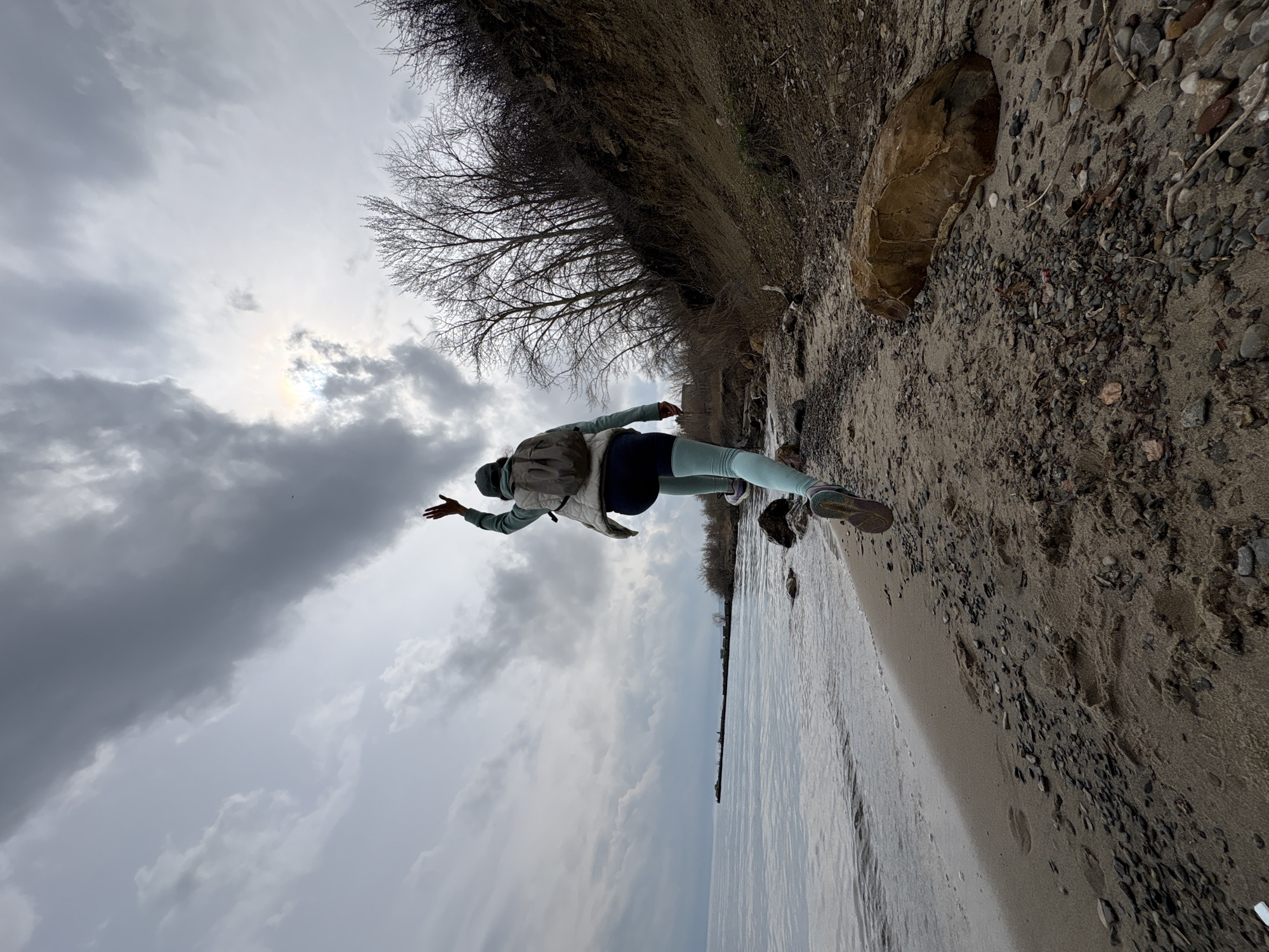 A woman stands on a sandy beach next to a body of water under a cloudy sky. The woman is wearing a light jacket, dark leggings, and sneakers, and she has a backpack on. She has her arms outstretched. The beach is covered with sand and small pebbles, and there is a grassy area or cliff behind the beach. The image is taken outdoors on an overcast day. The location is identified as Ulyanovsk, Russia.