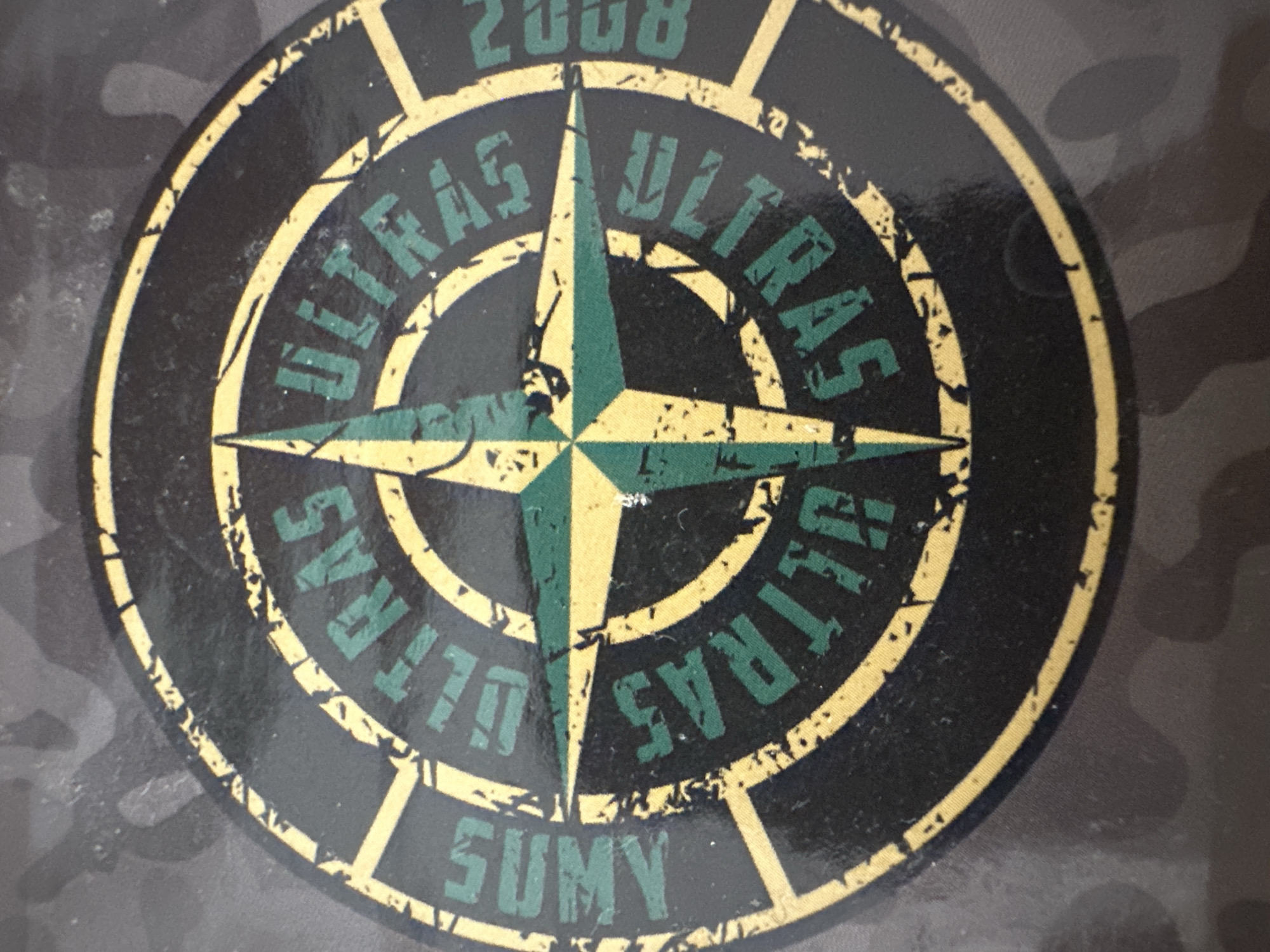 The image displays a close-up of a circular emblem with a distressed, vintage aesthetic. The emblem features a compass rose design with four points, rendered in green and yellow against a black background. The word "ULTRAS" is prominently displayed in green, encircling the compass rose at cardinal points. Between these, other words appear in green: "SUMY" on the left and "2008" on the right, oriented vertically. The emblem itself is set against a dark, camouflage-patterned fabric, likely clothing. The lighting is direct, highlighting the texture and wear of the emblem. The scene is static, with no visible people or actions. The location in Bečići, Montenegro, is not determinable from the visual information provided.